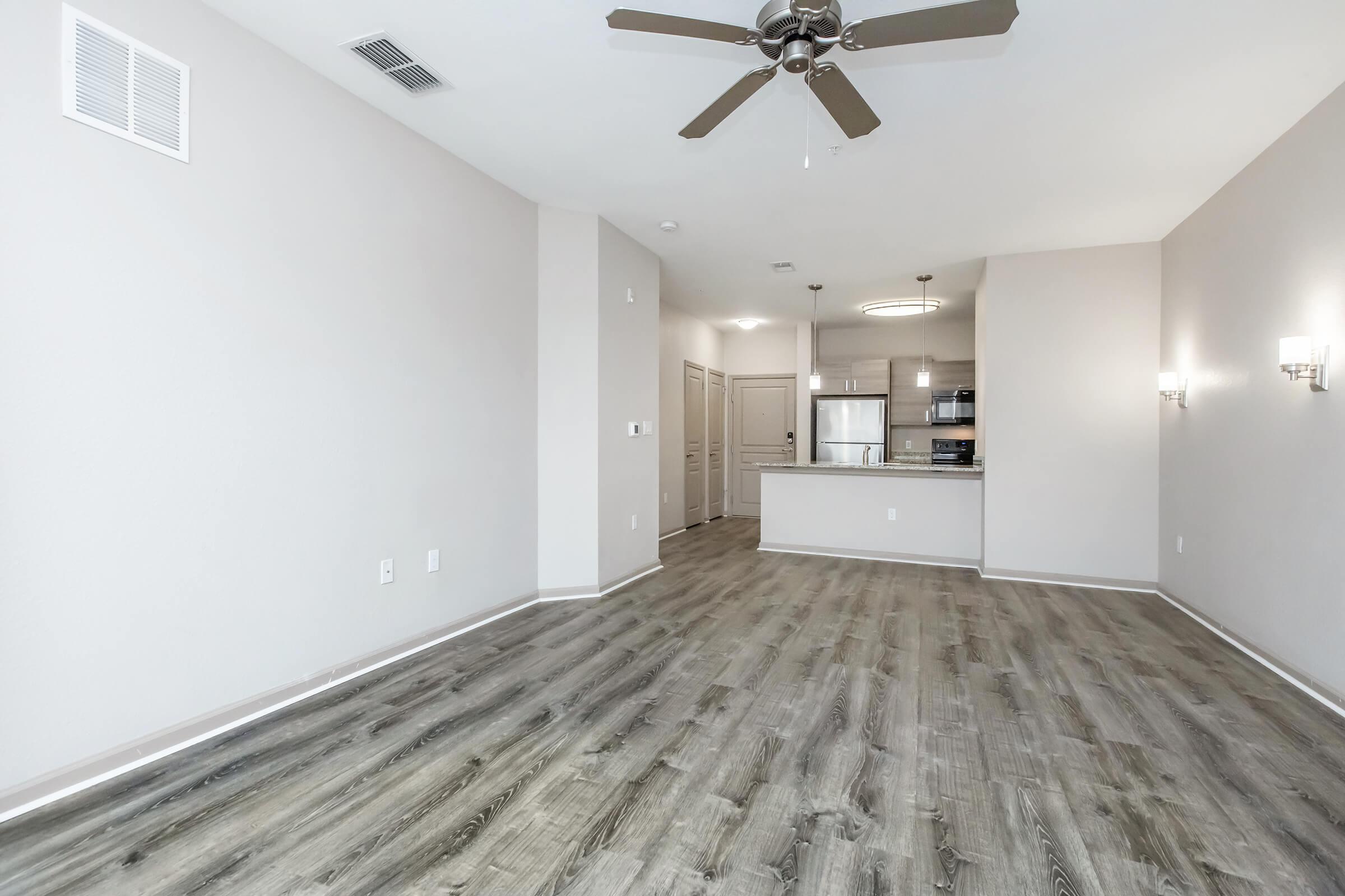 A spacious, well-lit living area with light gray walls and wood-style laminate flooring. The room features a ceiling fan and is connected to a kitchen area with modern appliances visible in the background. The space appears clean and ready for furnishing, with two sconce lights on the walls.