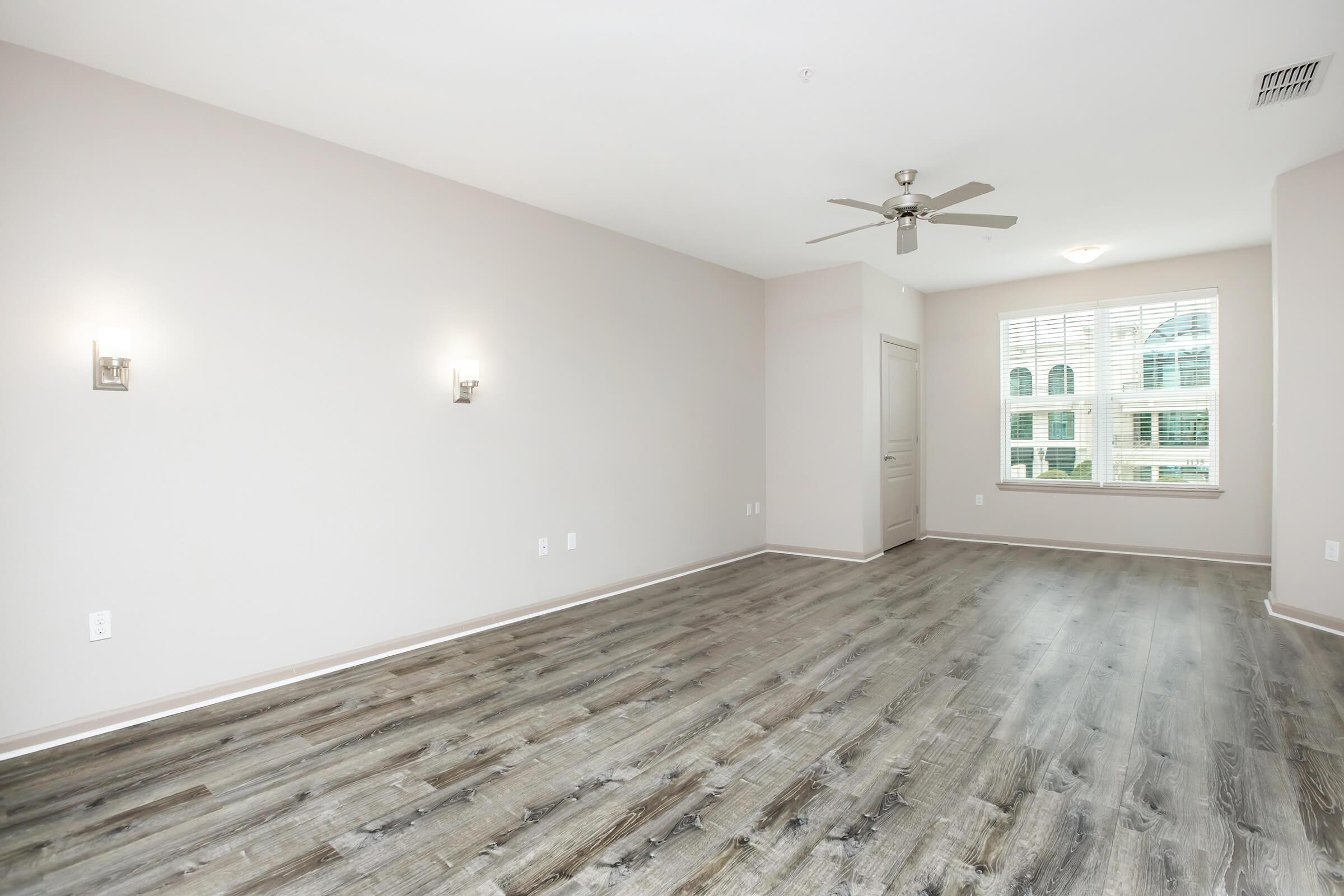 A spacious, empty room with light-colored walls and a ceiling fan. The floor is finished with a wood-like texture. Two wall sconces provide lighting, and there is a window with vertical blinds, allowing natural light to enter. The overall ambiance is bright and airy, ideal for personalization.
