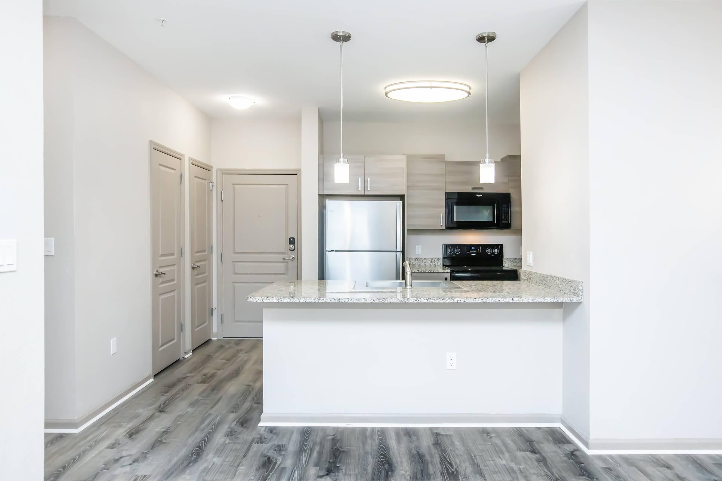 Modern kitchen and living area featuring gray cabinetry, stainless steel appliances, and a granite countertop. Includes pendant lighting over the kitchen island, light-colored walls, and laminate flooring. The entrance is visible with a door leading to a hallway.