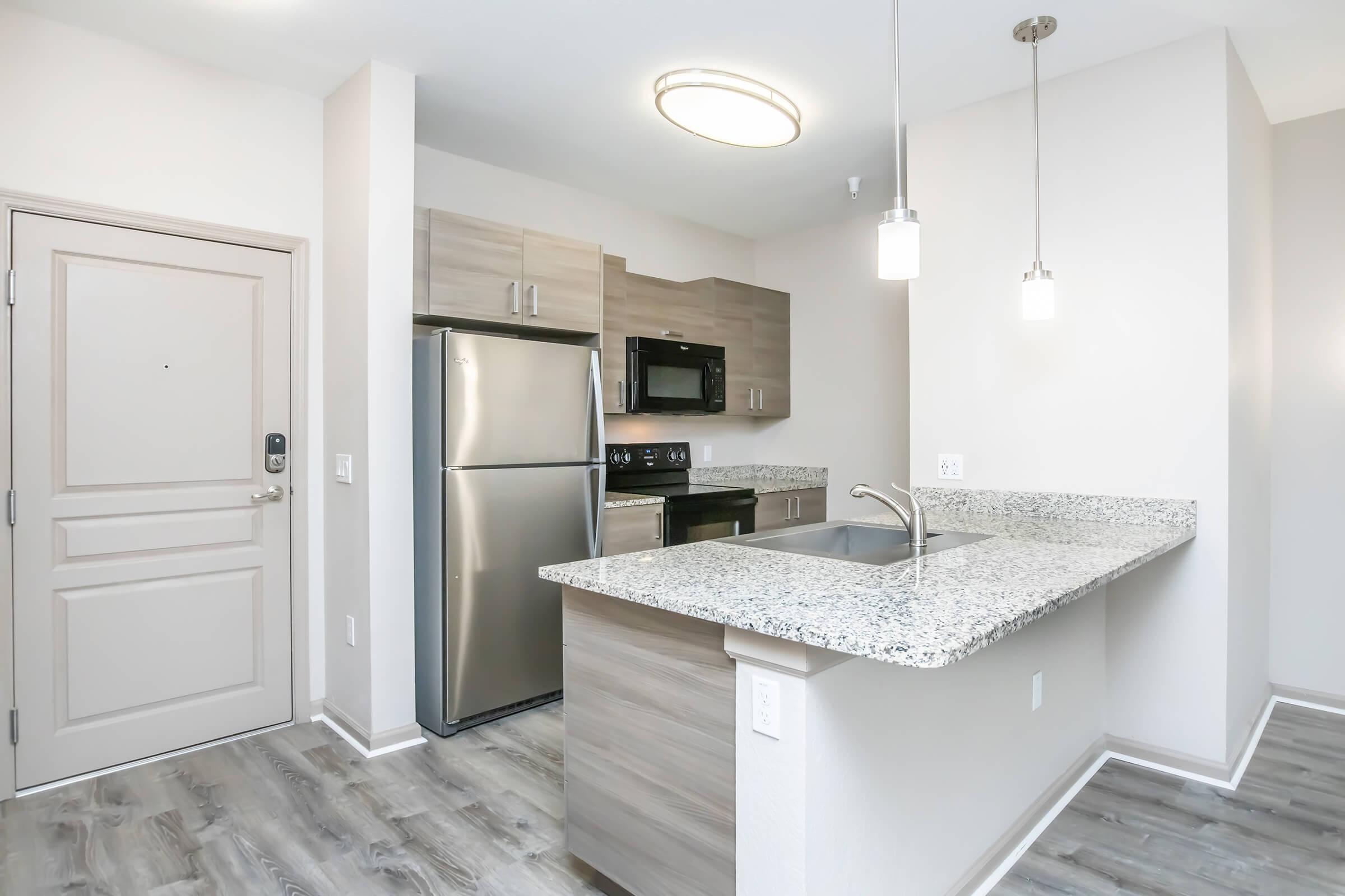 A modern kitchen featuring stainless steel appliances, including a refrigerator, microwave, and oven. The countertops are speckled granite, and there's a kitchen island with a sink. The space has light-colored walls and wood-style flooring, with a view of the entrance door. Pendant lights hang above the island.