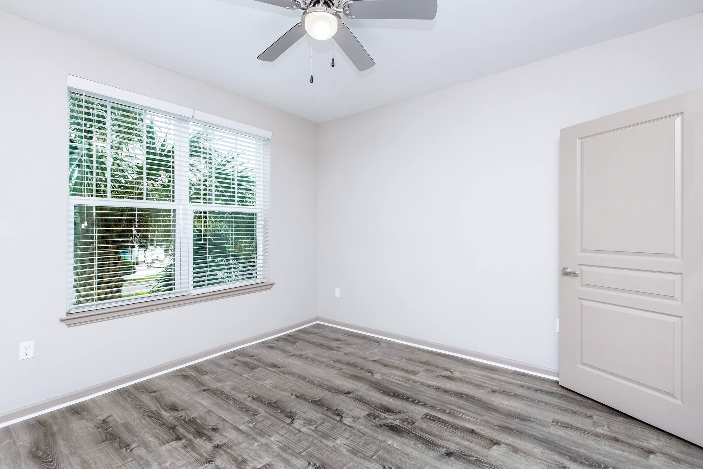 A well-lit, empty bedroom featuring a ceiling fan, large window with white blinds, light-colored walls, and laminate flooring. The door is closed, and the space is clean and ready for personalization or furnishing. Natural light enters from the window, creating a bright atmosphere.