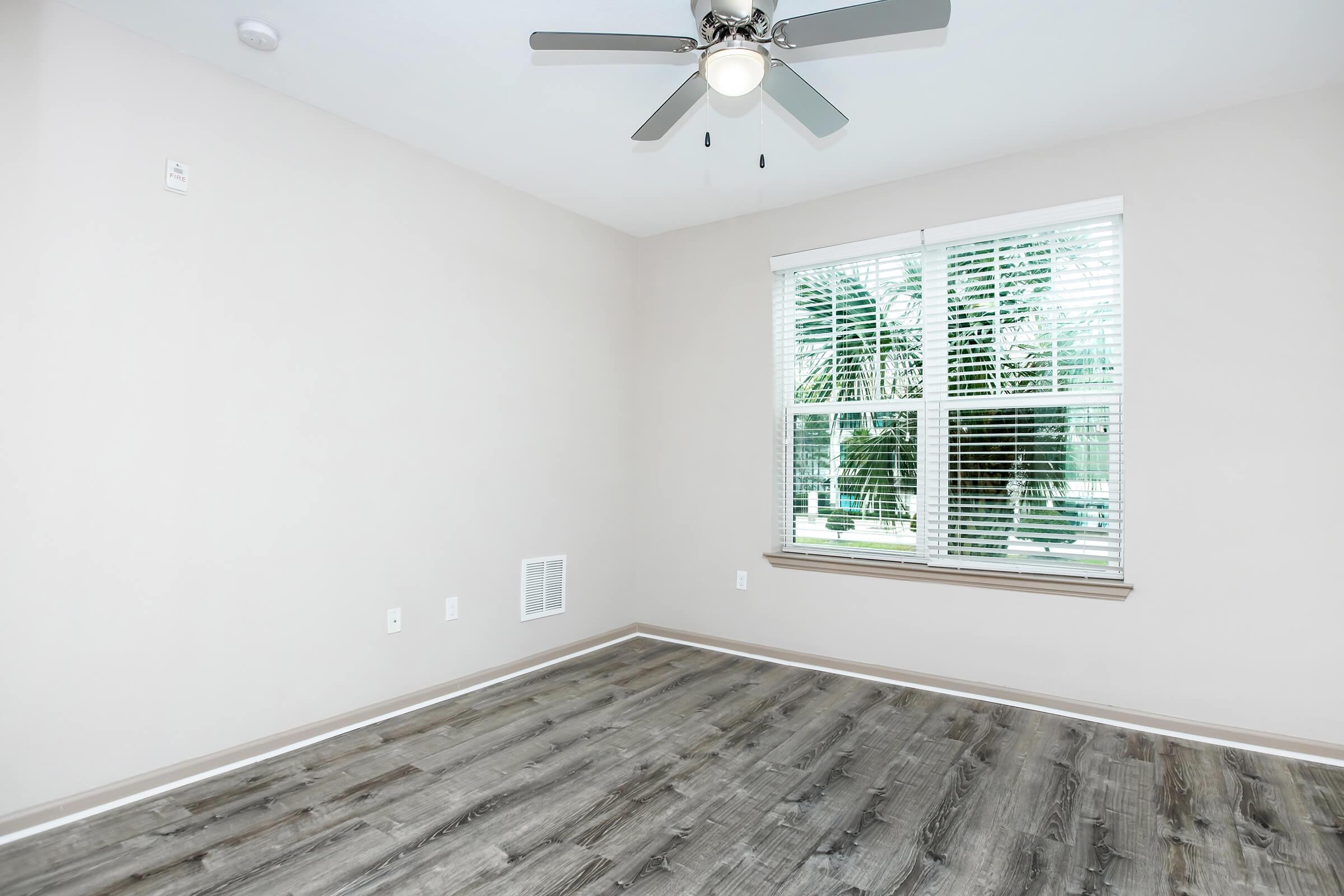 A bright, empty room with a light-colored wall and wooden flooring. There is a ceiling fan with light fixtures, and a window with blinds allowing natural light to enter. Outside the window, greenery can be seen, adding a touch of nature to the space.