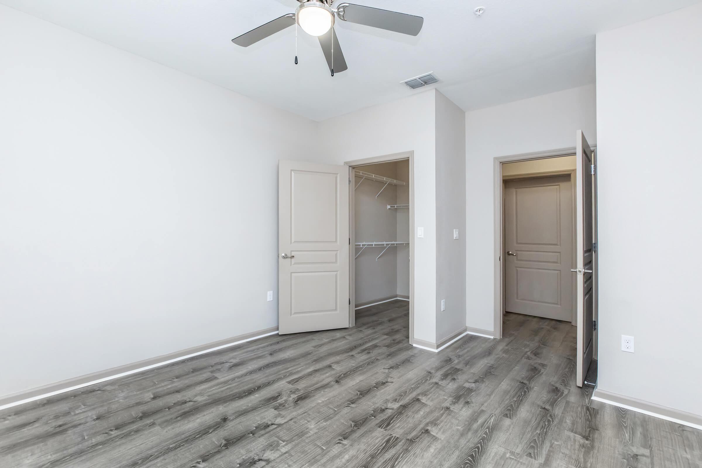 Empty room featuring a light-colored wall, a ceiling fan, and a light wood laminate floor. Two doorways lead to closets and other spaces, enhancing the room's layout. The overall atmosphere is bright and inviting, suitable for various uses such as a bedroom or office.