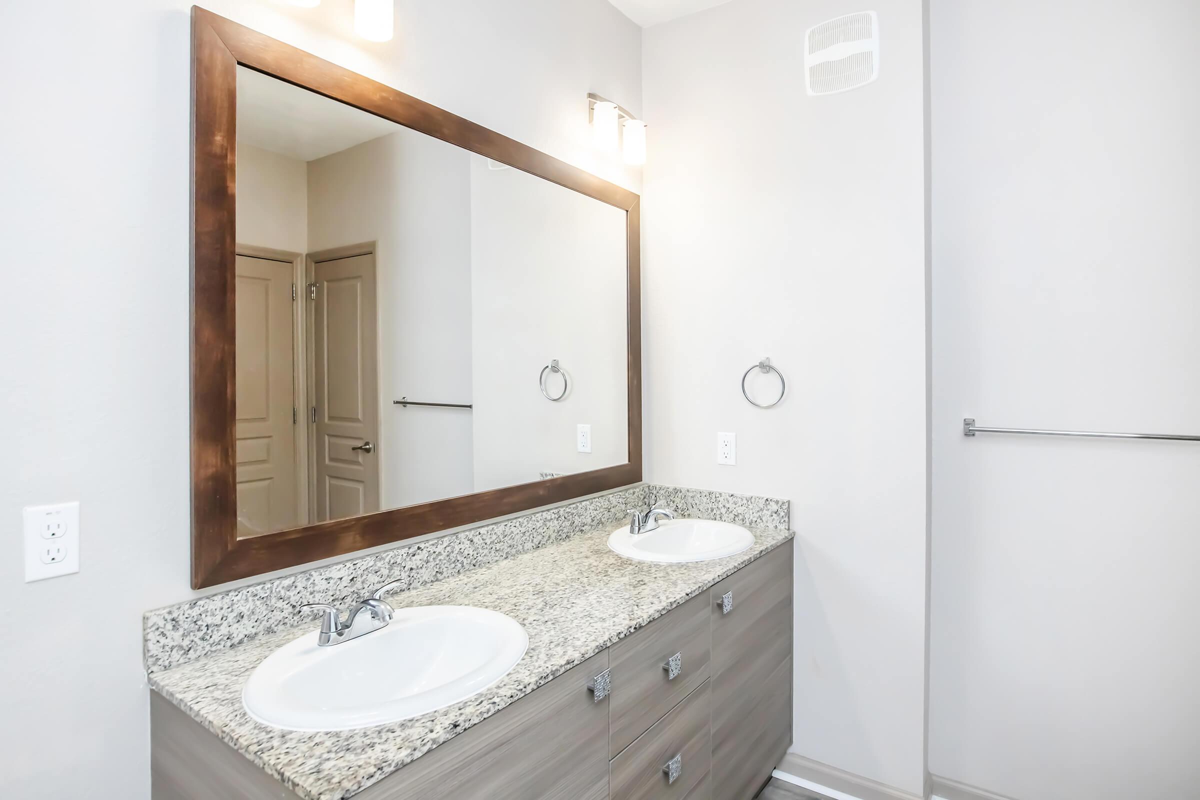 Modern bathroom featuring a double vanity with granite countertop, two sinks, and a large framed mirror. Soft gray walls complement the contemporary design, and there is a towel ring and towel bar for added convenience. The space is well-lit with overhead lighting.