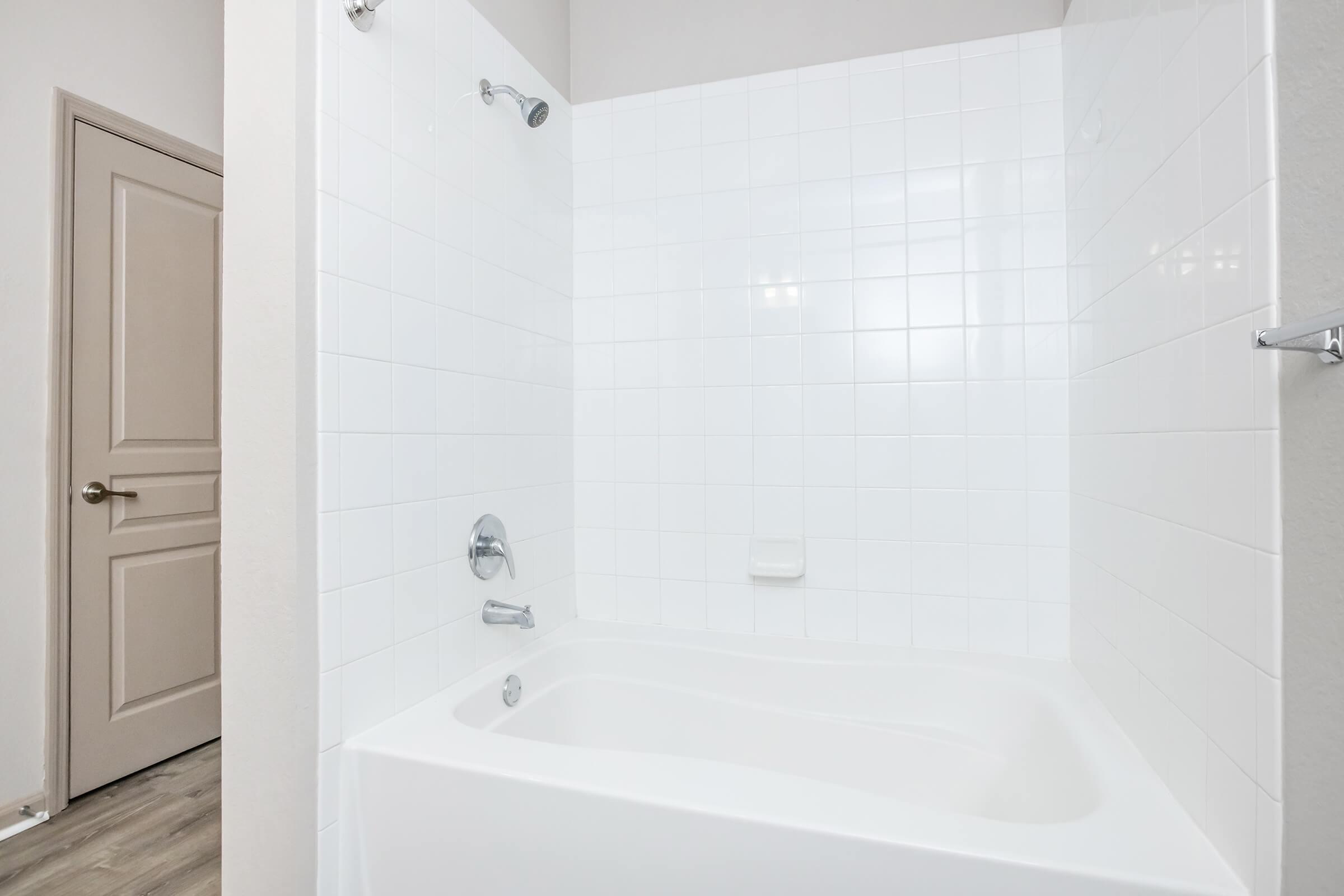 A clean and modern bathroom featuring a white tiled shower and bathtub. The shower has two showerheads, and the bathtub has a sleek design. A plain door is visible in the background, contributing to the minimalist aesthetic of the space.