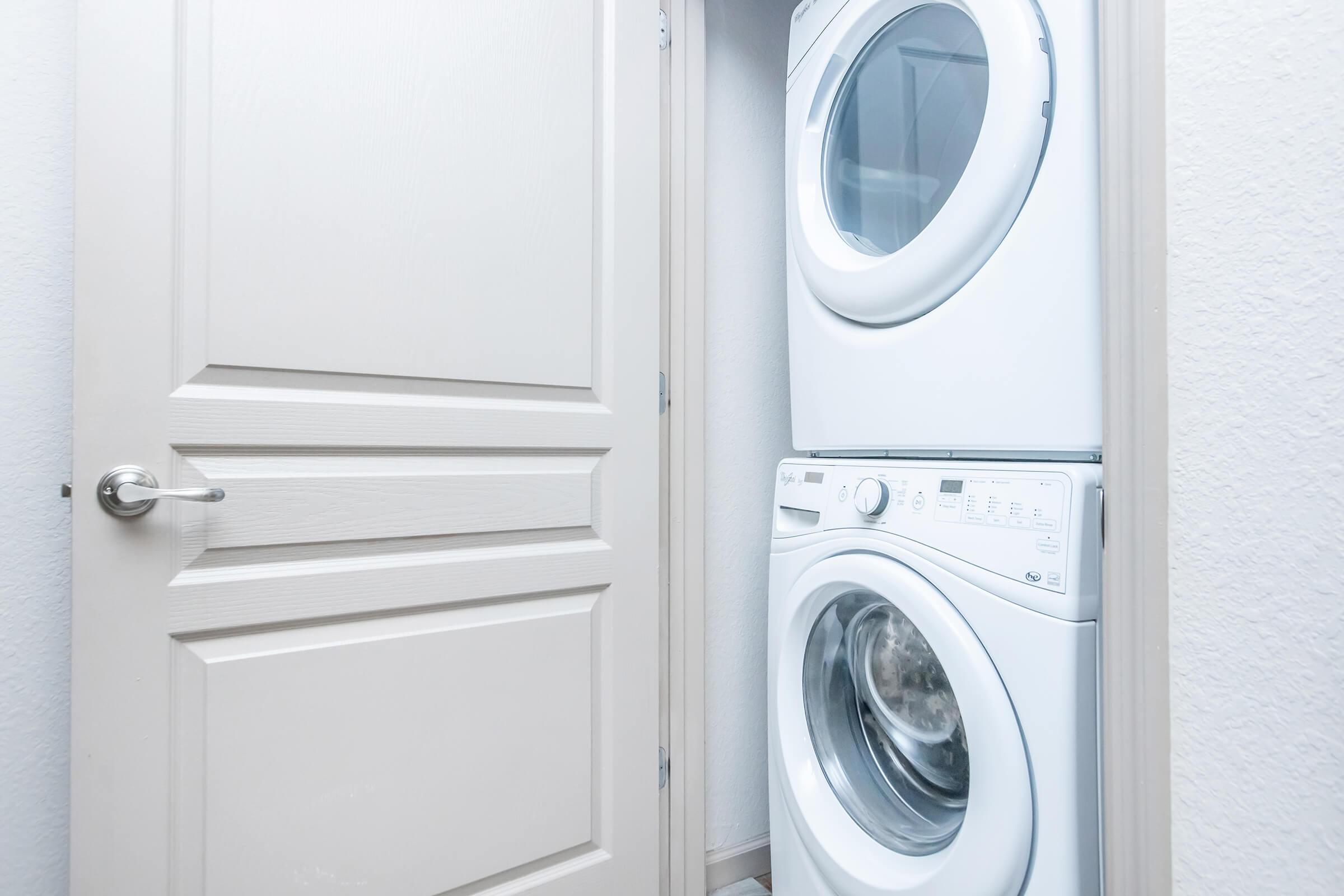 A stacked washer and dryer unit in a narrow laundry space, beside a closed light-colored door. The walls are painted in a neutral tone, and the overall setting appears clean and modern.