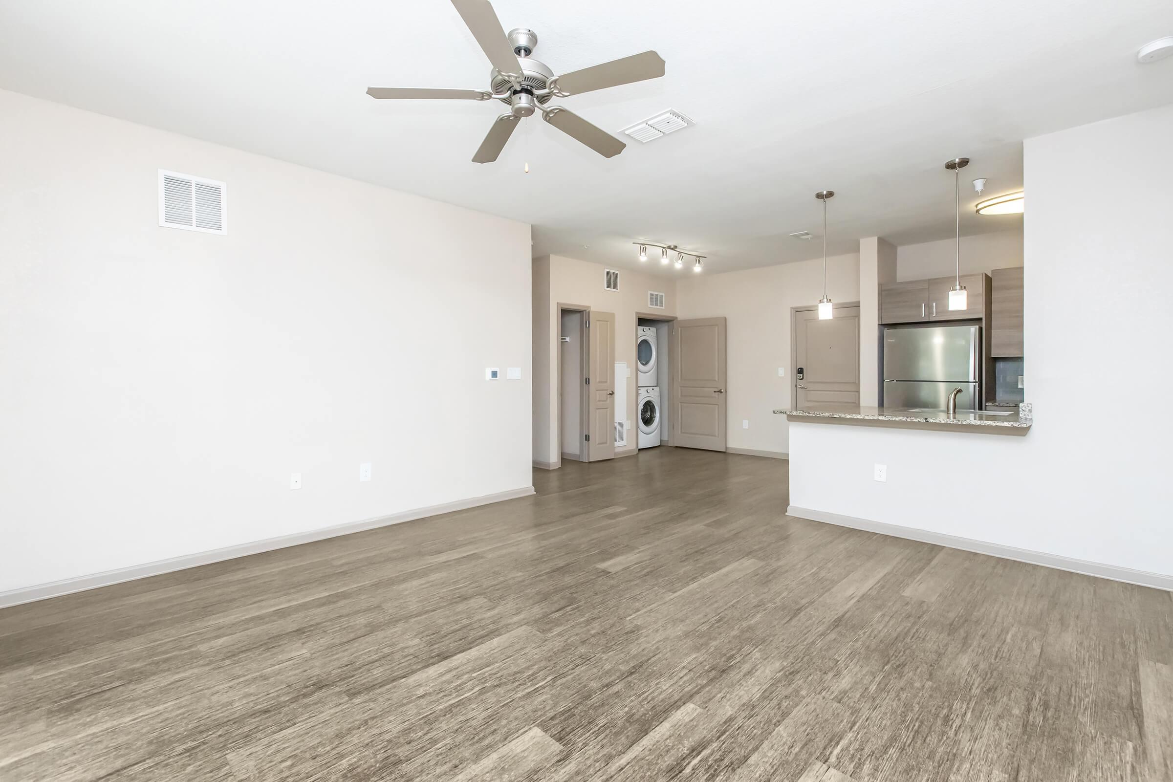 A spacious, modern living area featuring light-colored walls and hardwood-style flooring. A ceiling fan hangs above, and there's a kitchen visible with stainless steel appliances. The layout includes an area for a dining counter and washing machine, creating an open and inviting atmosphere.