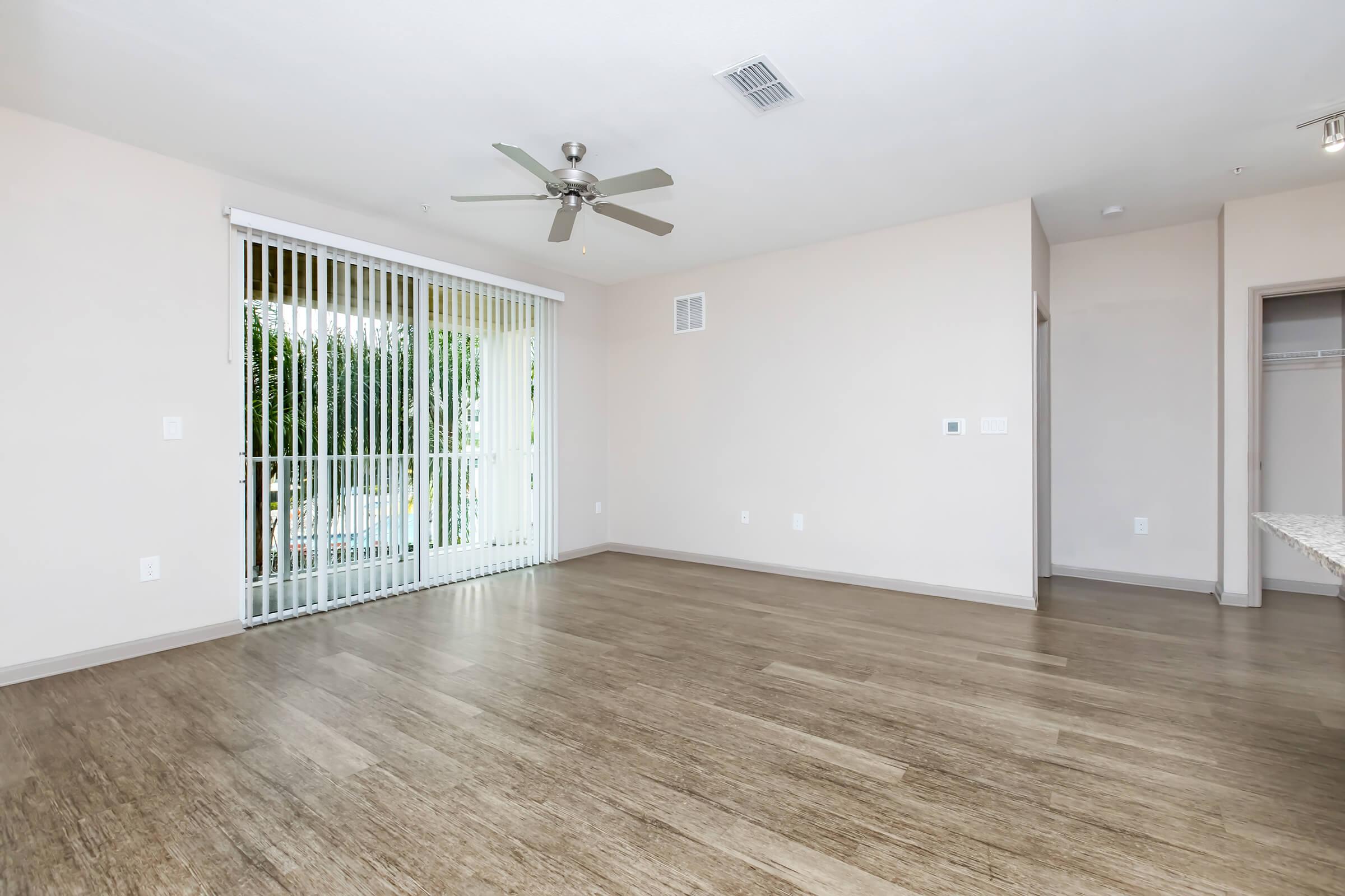 Bright, spacious interior of a living room featuring light-colored walls, wooden flooring, and a ceiling fan. Large sliding glass doors with vertical blinds lead to an outdoor area. A closet is visible on the right side, creating a cozy, inviting atmosphere.