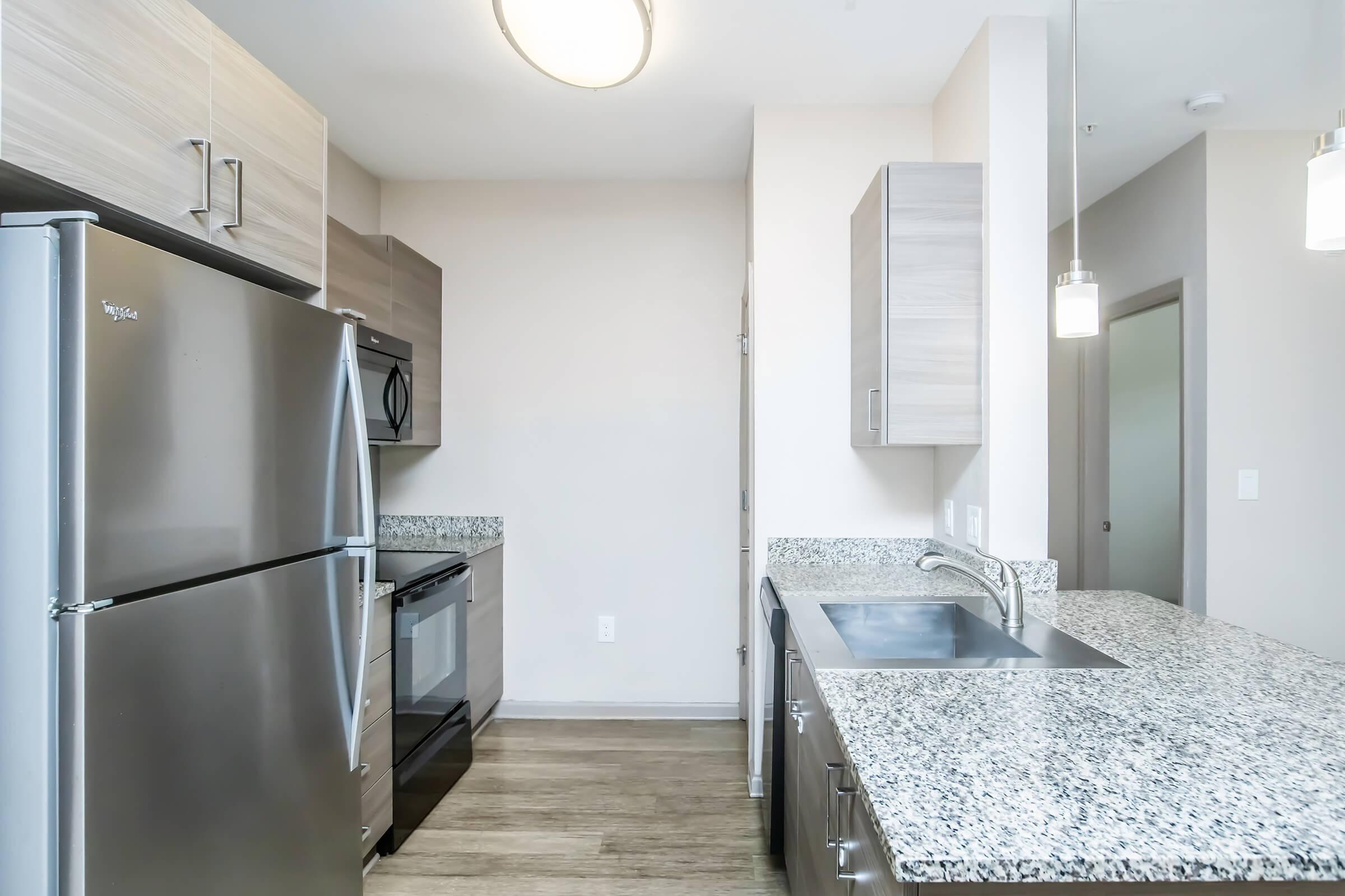 Modern kitchen featuring stainless steel appliances, including a refrigerator and oven. The countertops are made of light-colored granite, and the cabinetry has a sleek design. Bright lighting fixtures hang from the ceiling, complementing the neutral color scheme of the walls and flooring.