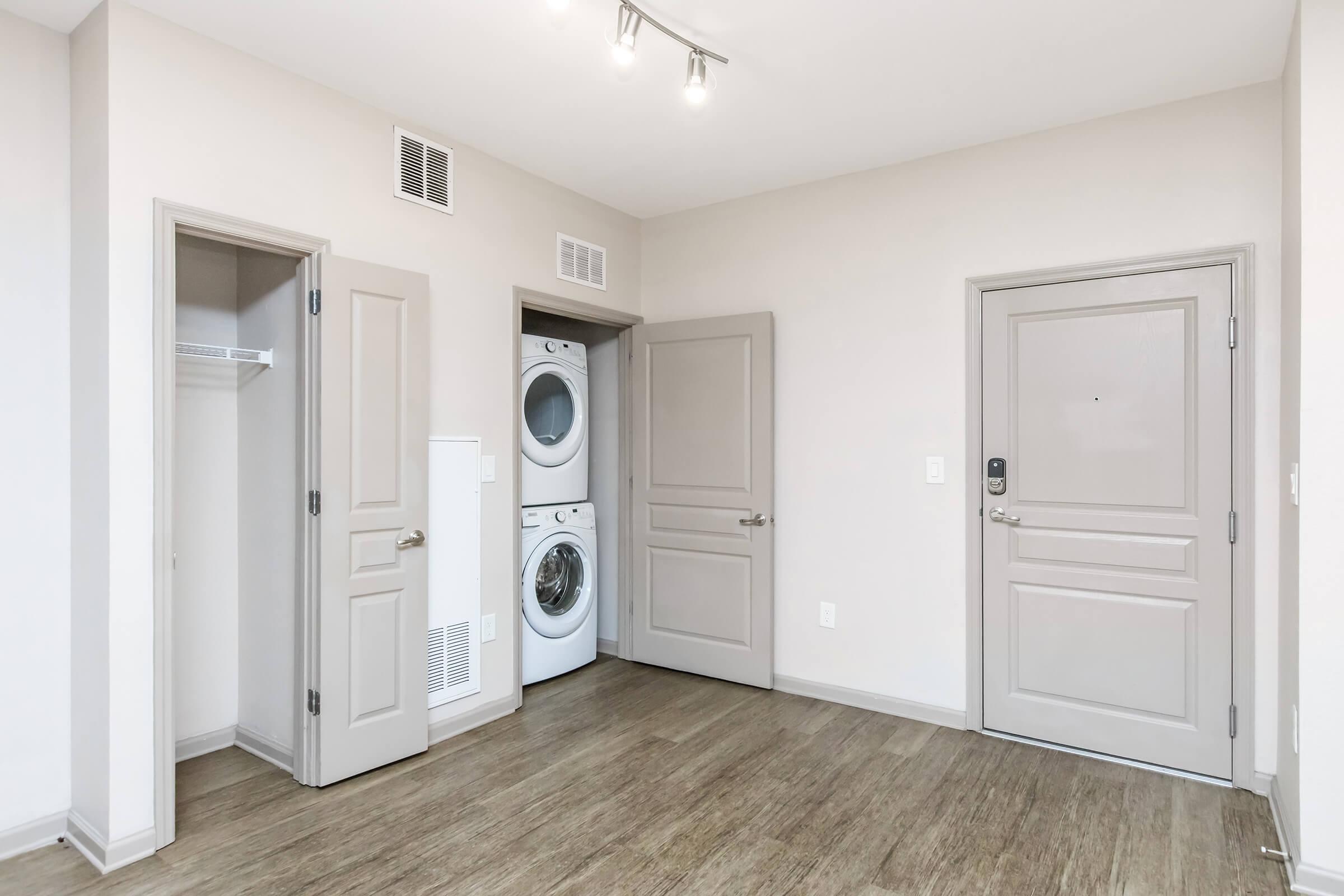A small laundry area in a modern apartment, featuring a stacked washer and dryer. There are two closed doors, one leading to a closet with a shelf, and the other to the main entrance. The walls are a light color, and the flooring is a wood-like laminate. Ceiling lights provide bright illumination.