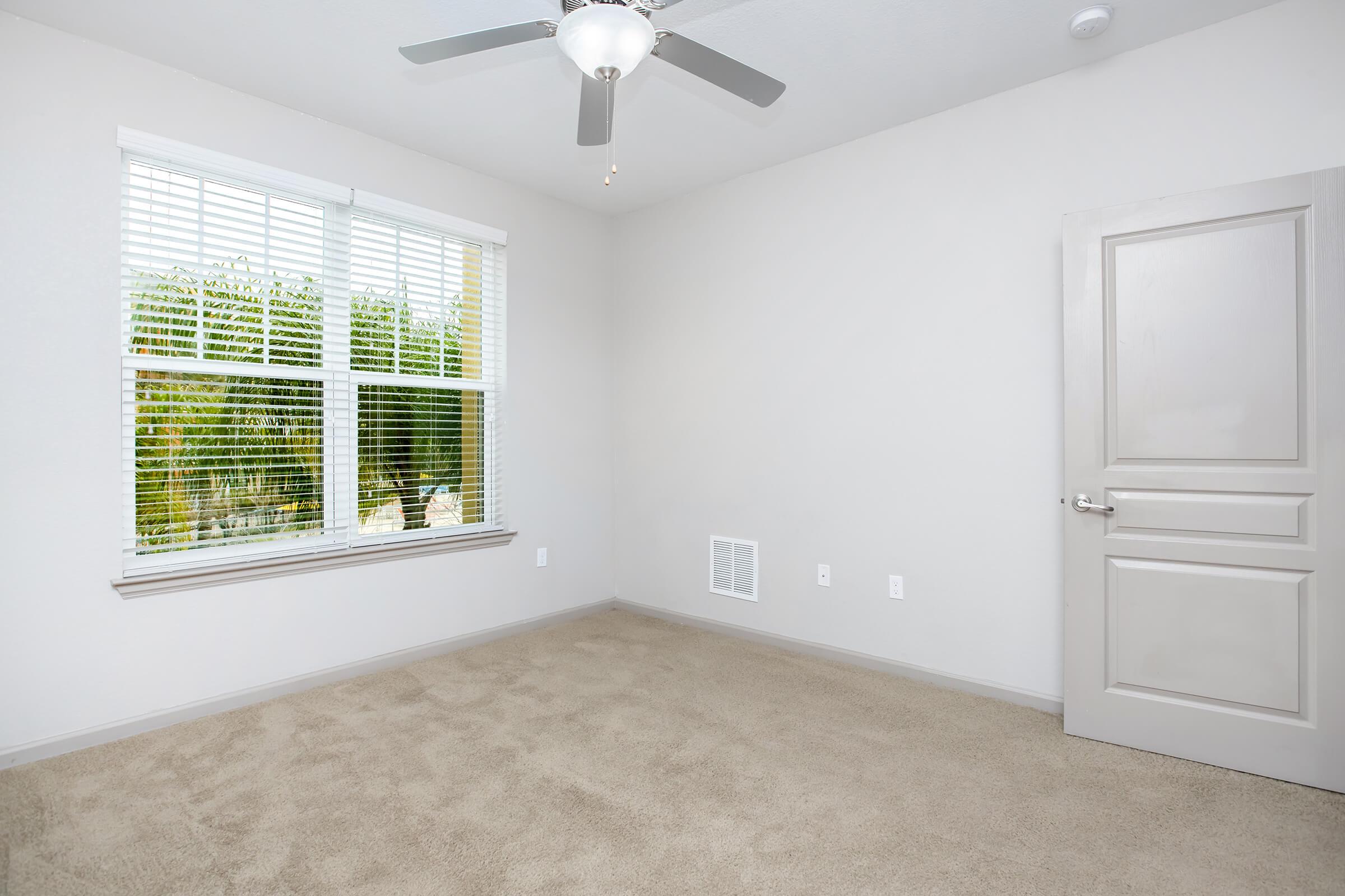 A well-lit, sparsely furnished room featuring beige carpet, a ceiling fan, and a large window with white blinds. The walls are painted a light gray color, and there’s a closed door on one side. Green foliage can be seen outside the window, adding a touch of nature to the scene.