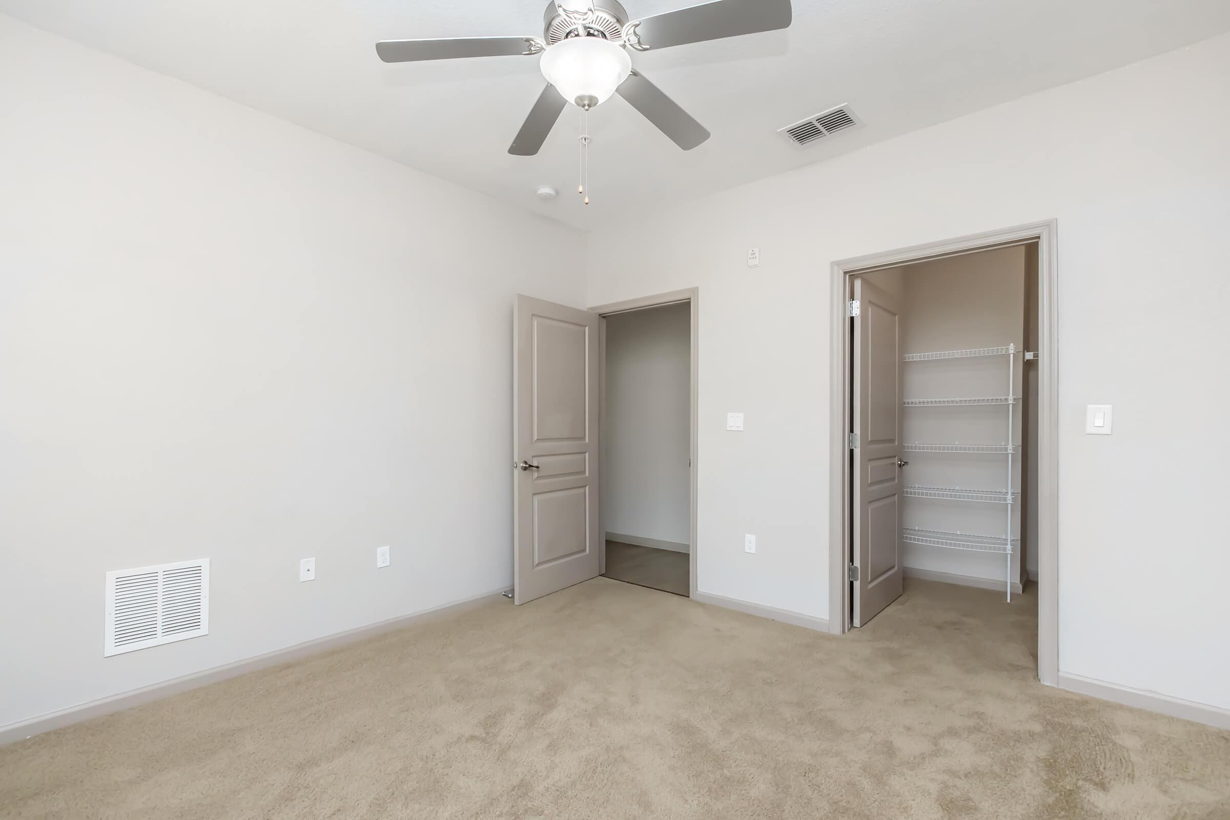 A well-lit, neutral-toned bedroom featuring beige carpet, a ceiling fan, and a closed door leading to a closet. An open door reveals another room, and the walls are painted light gray. The space is unfurnished, creating an airy and open feel.