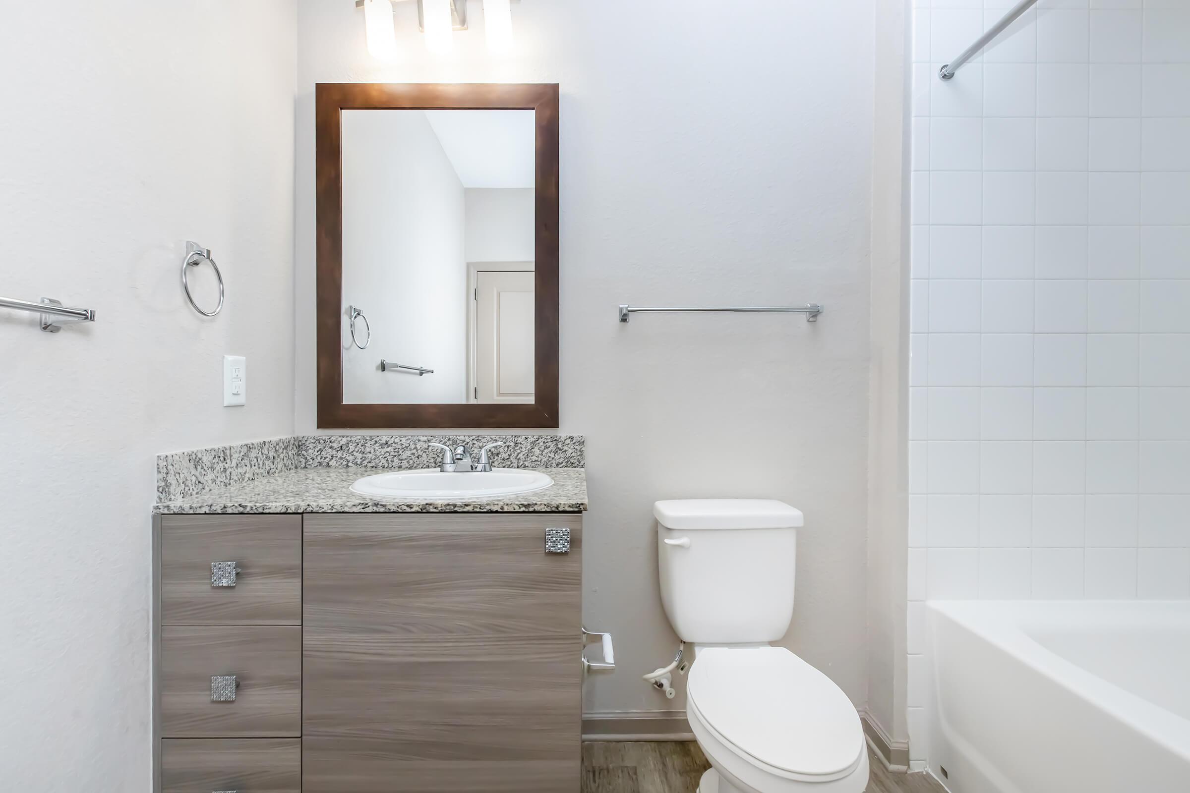 A modern bathroom featuring a granite countertop vanity with a rectangular mirror above, a white toilet, and a bathtub with a shower rail. The walls are light-colored, and there is a towel bar on the wall. The floor has a wood-like design, creating a clean and contemporary look.