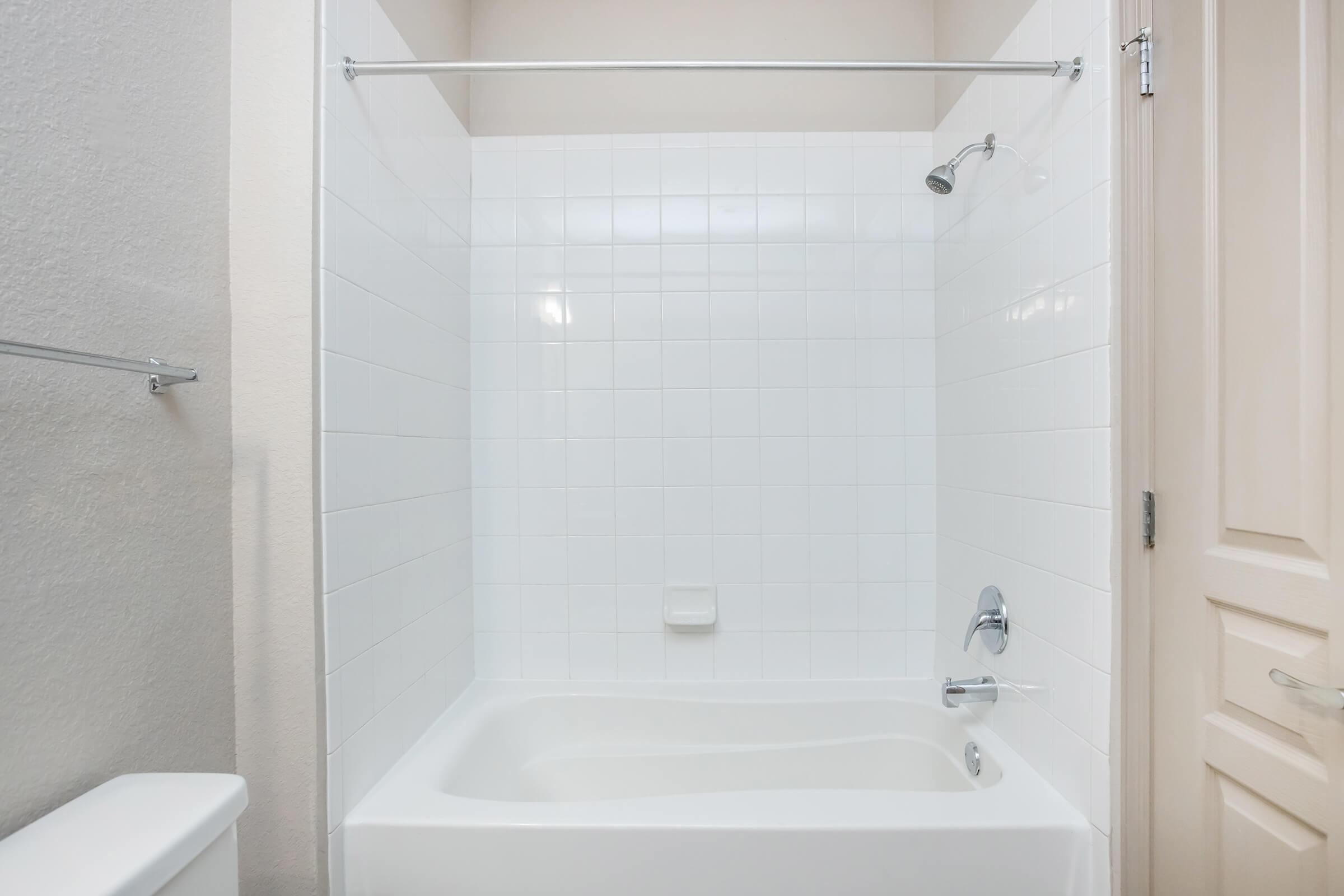 A clean, modern bathroom featuring a white tiled shower area with a tub. The showerhead is mounted on the wall, and there are minimal fixtures. The space is bright and uncluttered, with light-colored walls and a door visible in the background. A white toilet is also partially visible on the left.