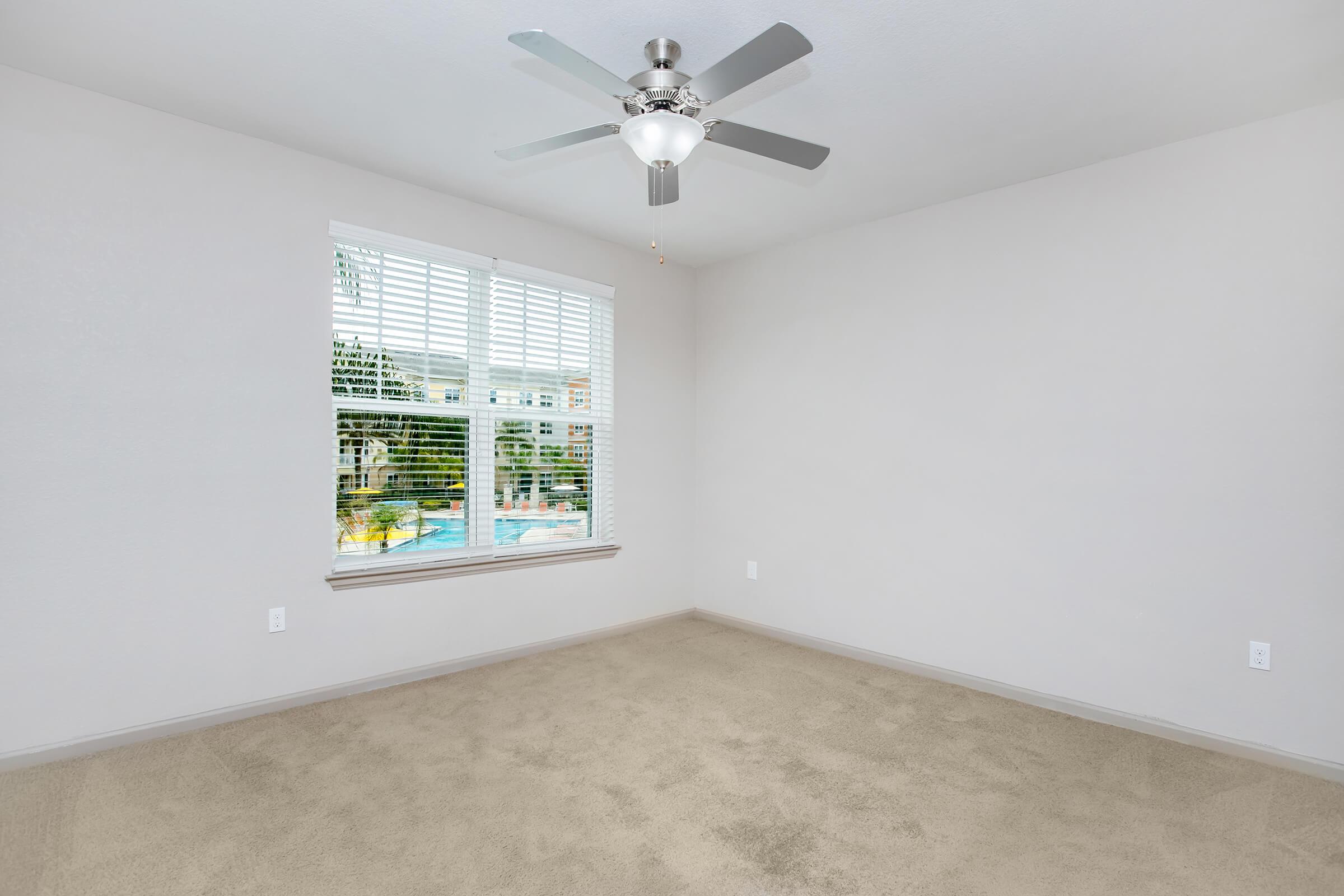Empty room with light beige carpet and gray walls. A ceiling fan with a light fixture is mounted on the ceiling. A window with white blinds allows natural light inside, revealing a view of a pool and greenery outside.