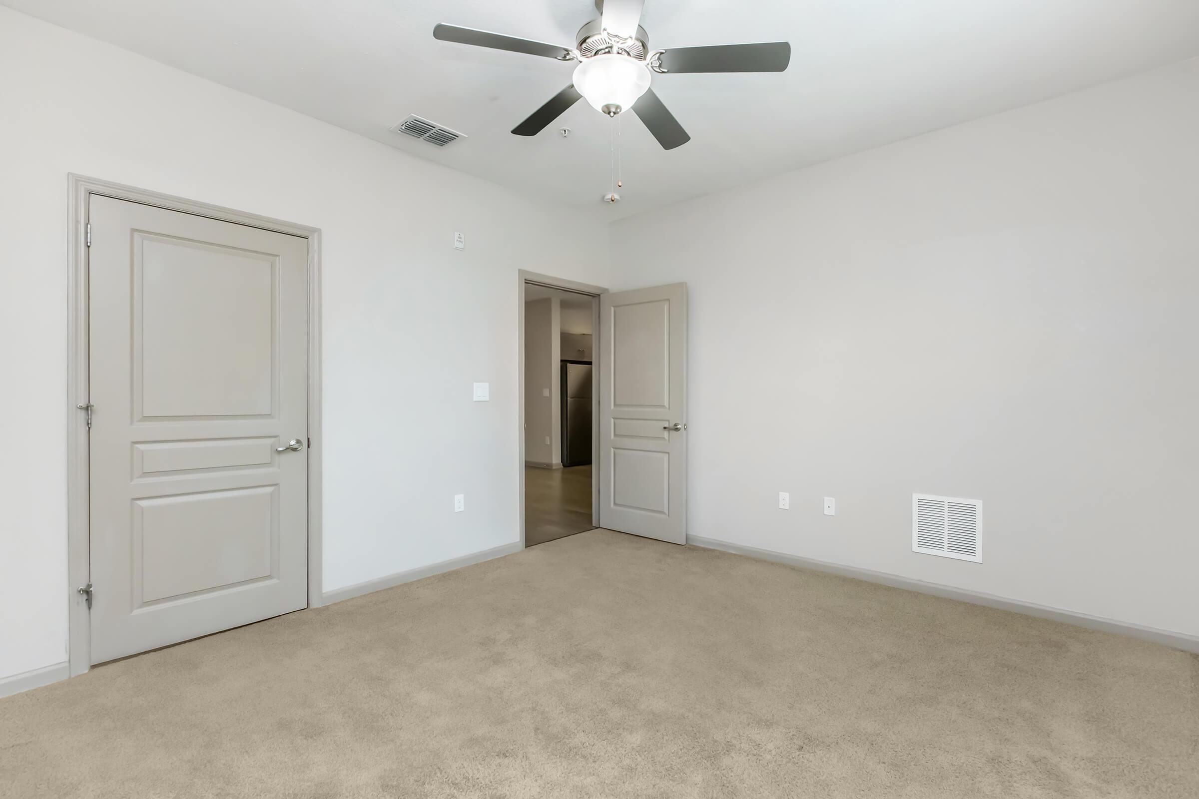 A spacious, empty room with light beige walls and carpet. There are two closed doors on the left side, and a ceiling fan with three blades hangs from the ceiling. A wall vent is visible at the bottom right, and the overall lighting is bright and neutral, creating an inviting atmosphere.