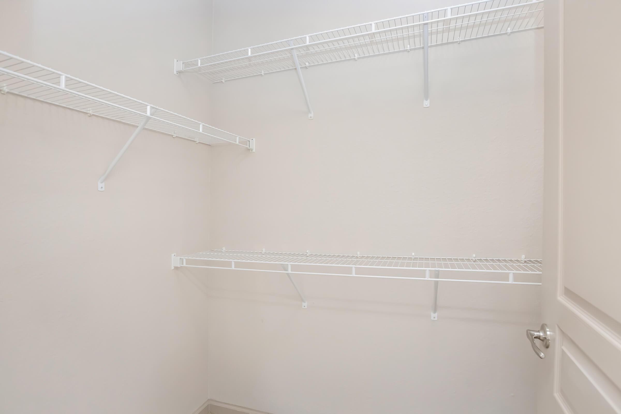 Empty closet with white wire shelving attached to light-colored walls. The shelves are positioned at different heights, providing storage space. The floor is bare and clean, creating a minimalistic look.