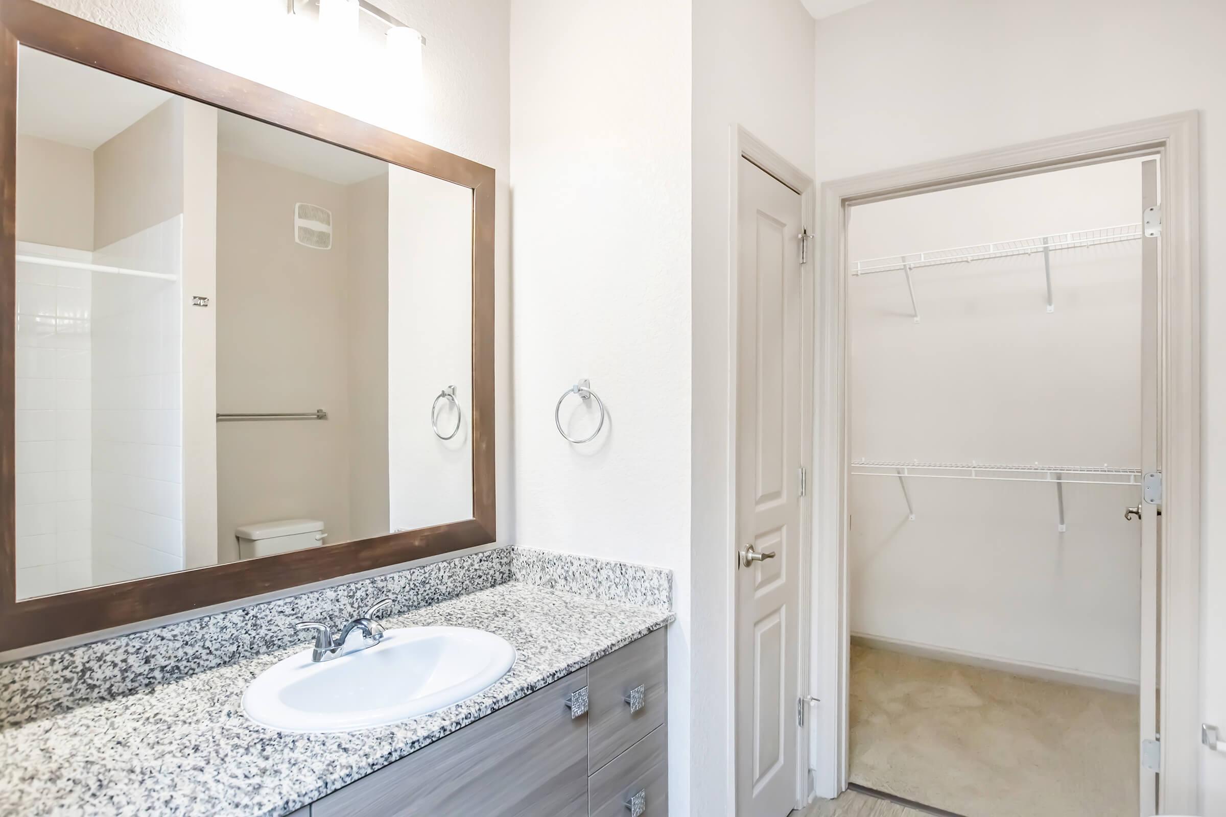 A modern bathroom with a large mirror above a granite countertop sink. It features a shower area with white tiles on one side and a doorway leading to an empty walk-in closet on the right. The walls are painted in a light beige shade, creating a bright and spacious atmosphere.