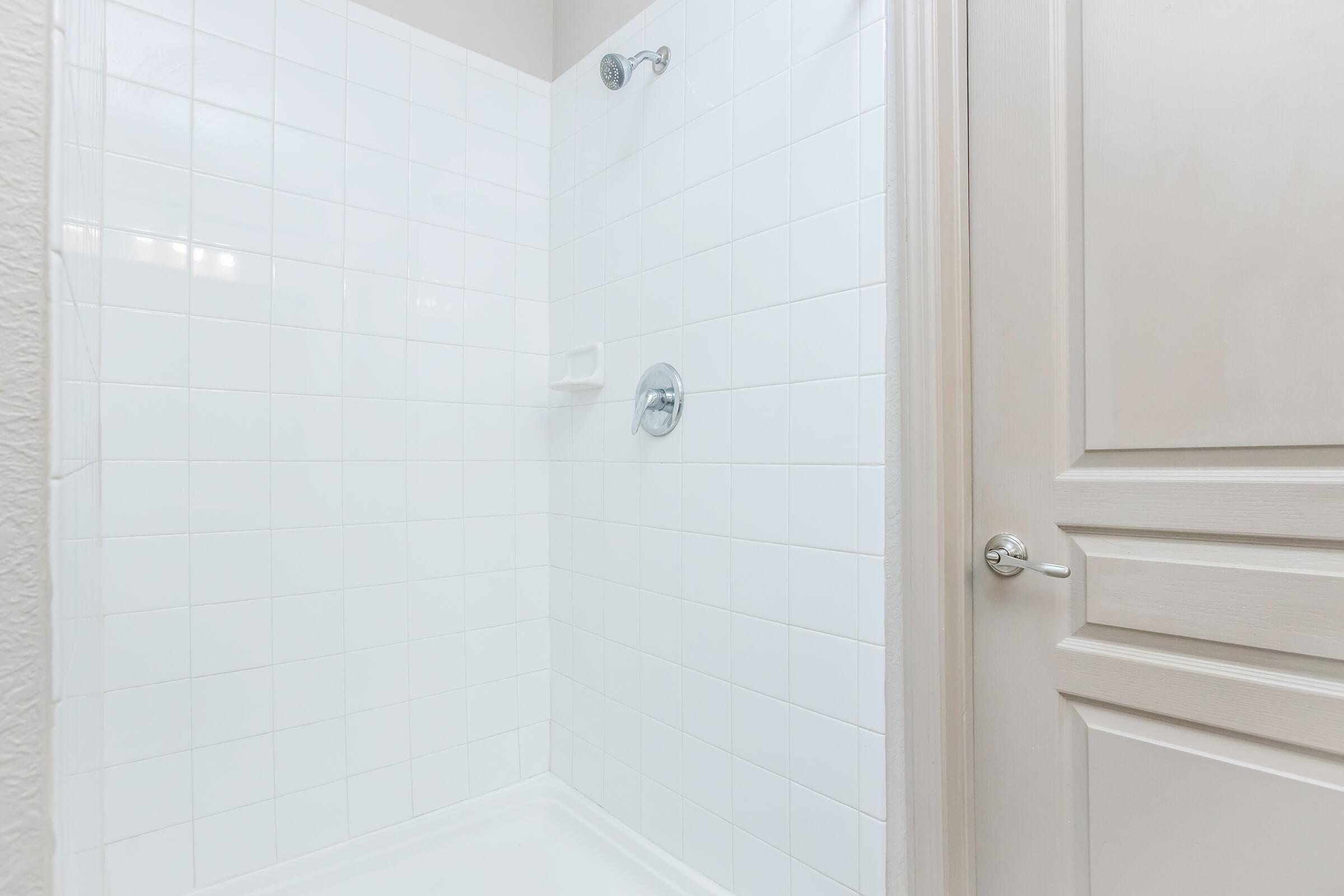 A clean and simple bathroom shower area with white tiled walls, a handheld showerhead, and a chrome faucet. There is a white corner shelf and a closed wooden door with a silver handle nearby. The space is well-lit, giving a bright and fresh appearance.