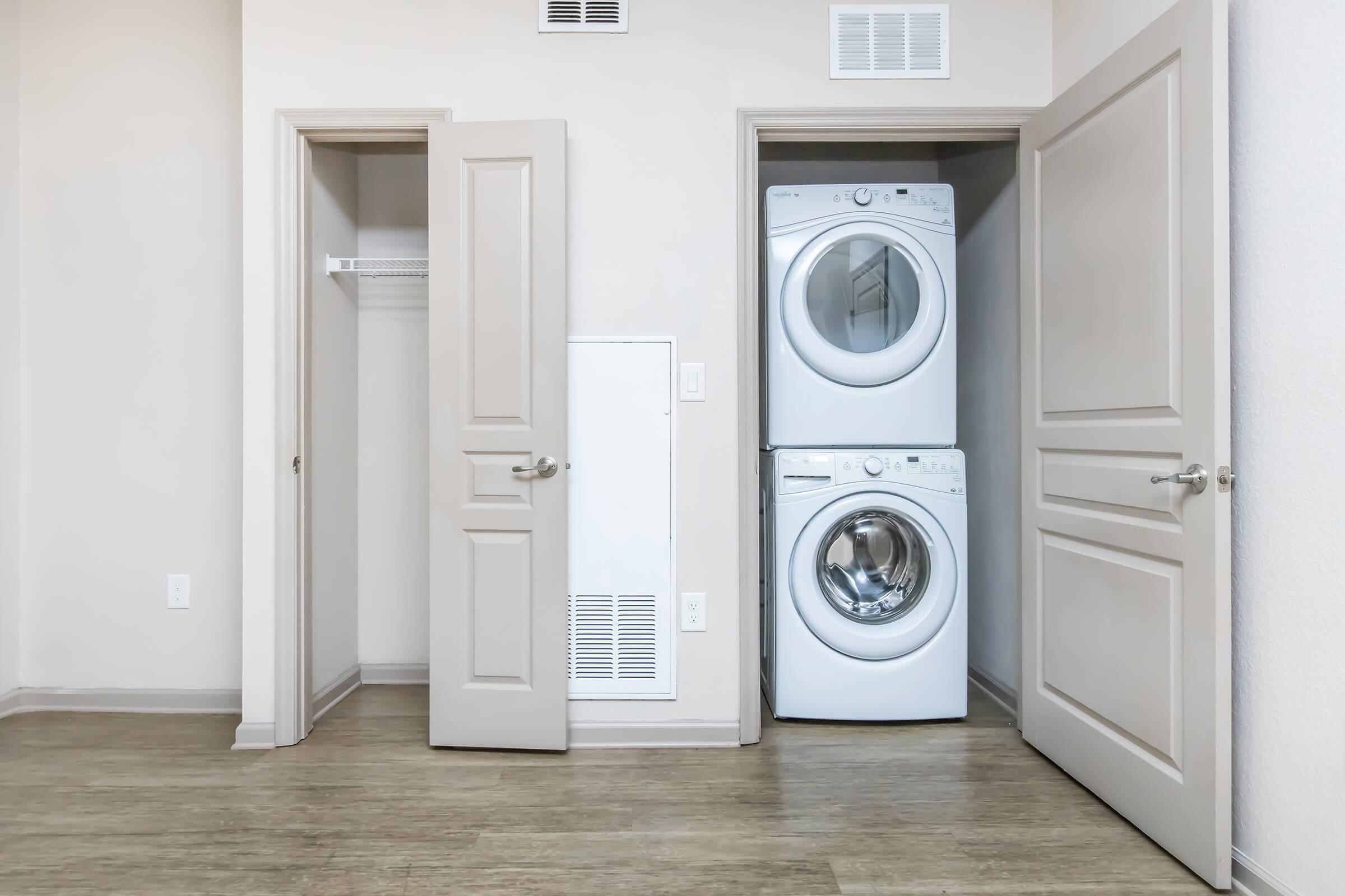 A laundry room featuring a stacked washer and dryer in an alcove, with two closet doors on either side. The walls are a neutral color, and the flooring is light wood. One door is slightly open, revealing a small shelf.