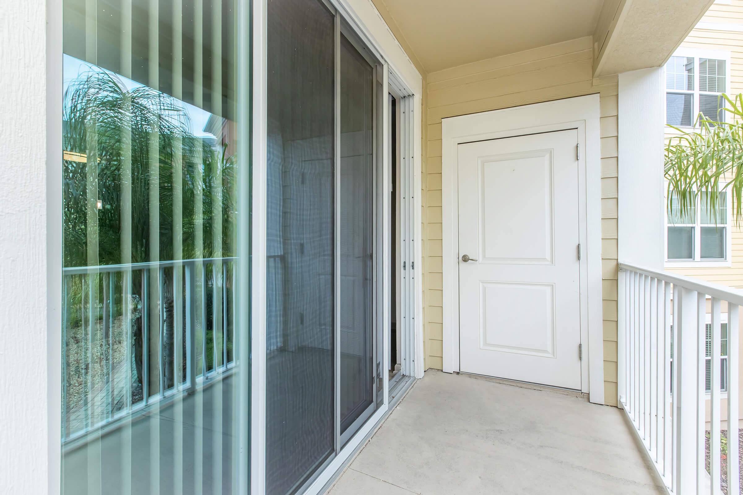 A view of a balcony featuring sliding glass doors with vertical blinds, leading to an entrance door. The exterior has a light-colored finish with beige siding, and plants are visible in the background, suggesting a residential setting.