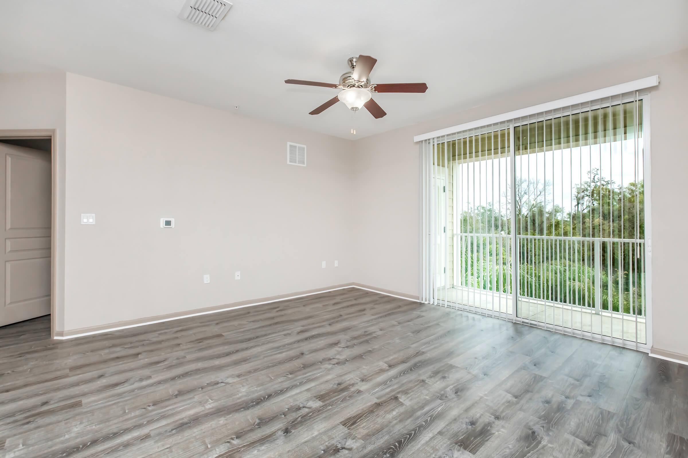 Spacious, empty living room with light-colored walls and wood-like flooring. A ceiling fan hangs from the ceiling, and large sliding glass doors lead to a balcony. Natural light floods the space, highlighting the open layout with no furniture present. Ideal for personalization and design.