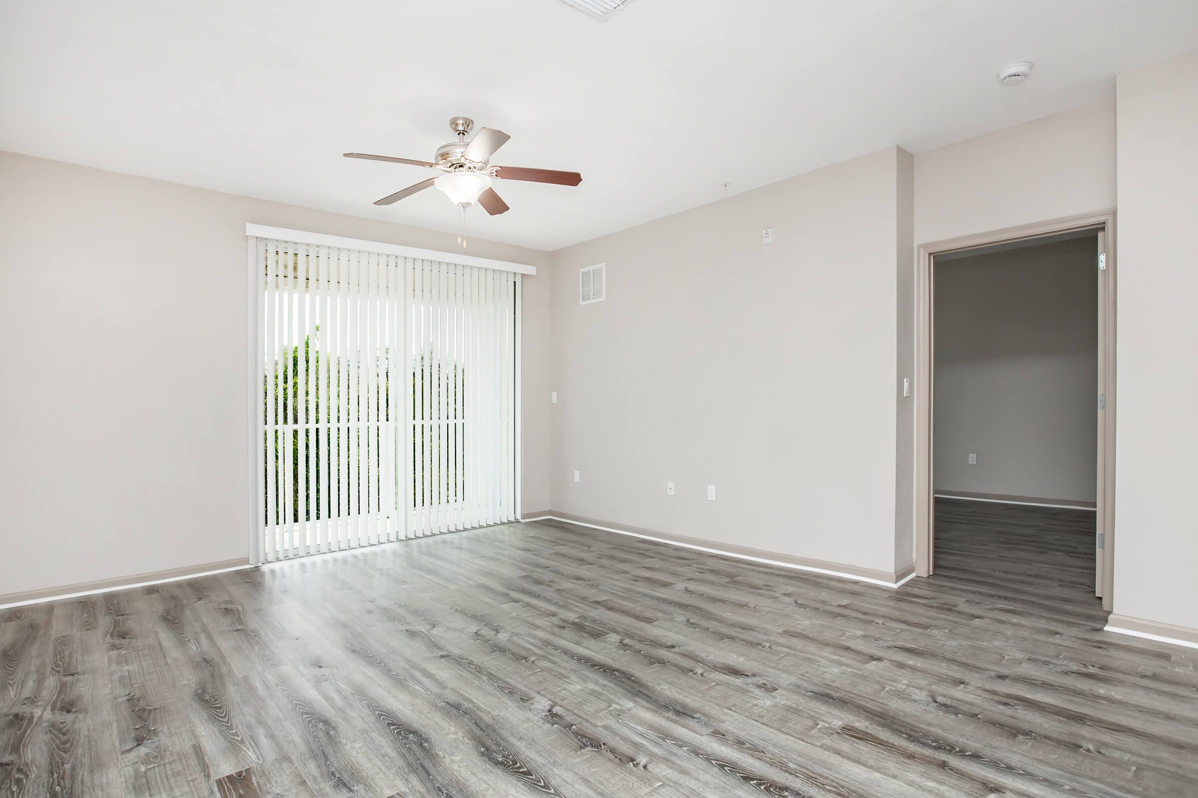 Spacious interior of a modern living room featuring beige walls, wood-like laminate flooring, a ceiling fan, and vertical blinds covering a large window. A doorway to another room is visible on the right, and the overall ambiance is bright and inviting.