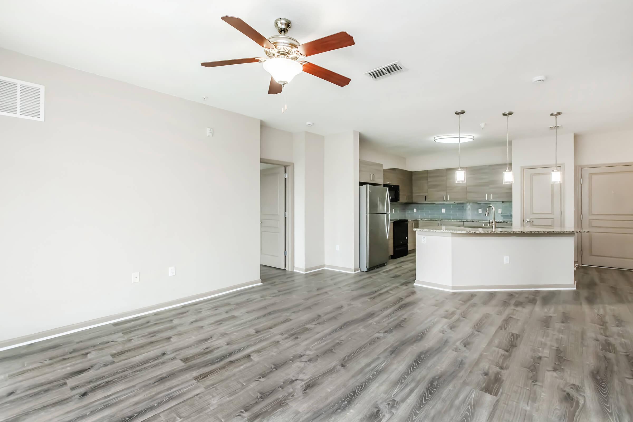 A spacious, modern living room with gray wood-style flooring, a ceiling fan, and light-colored walls. The open kitchen features stainless steel appliances, granite countertops, and pendant lighting. Abundant natural light fills the space, creating a welcoming atmosphere.