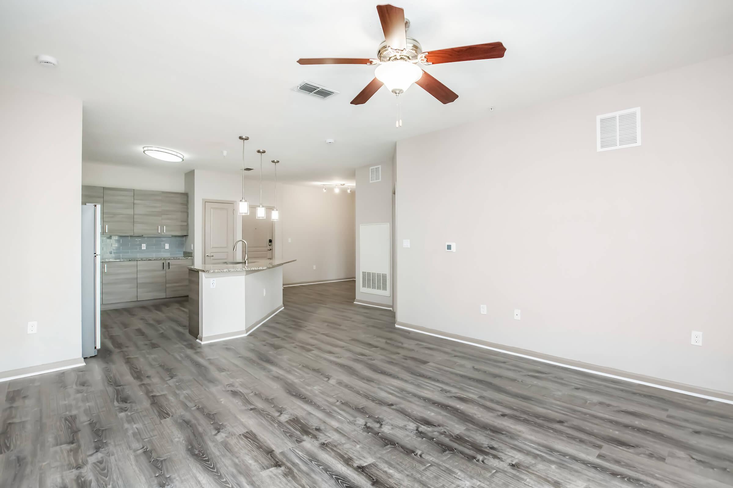 Spacious interior of a modern apartment featuring light-colored walls, wood-like flooring, and a ceiling fan. The open layout connects to a kitchen with gray cabinets and a countertop. Natural light illuminates the room, creating a welcoming atmosphere.