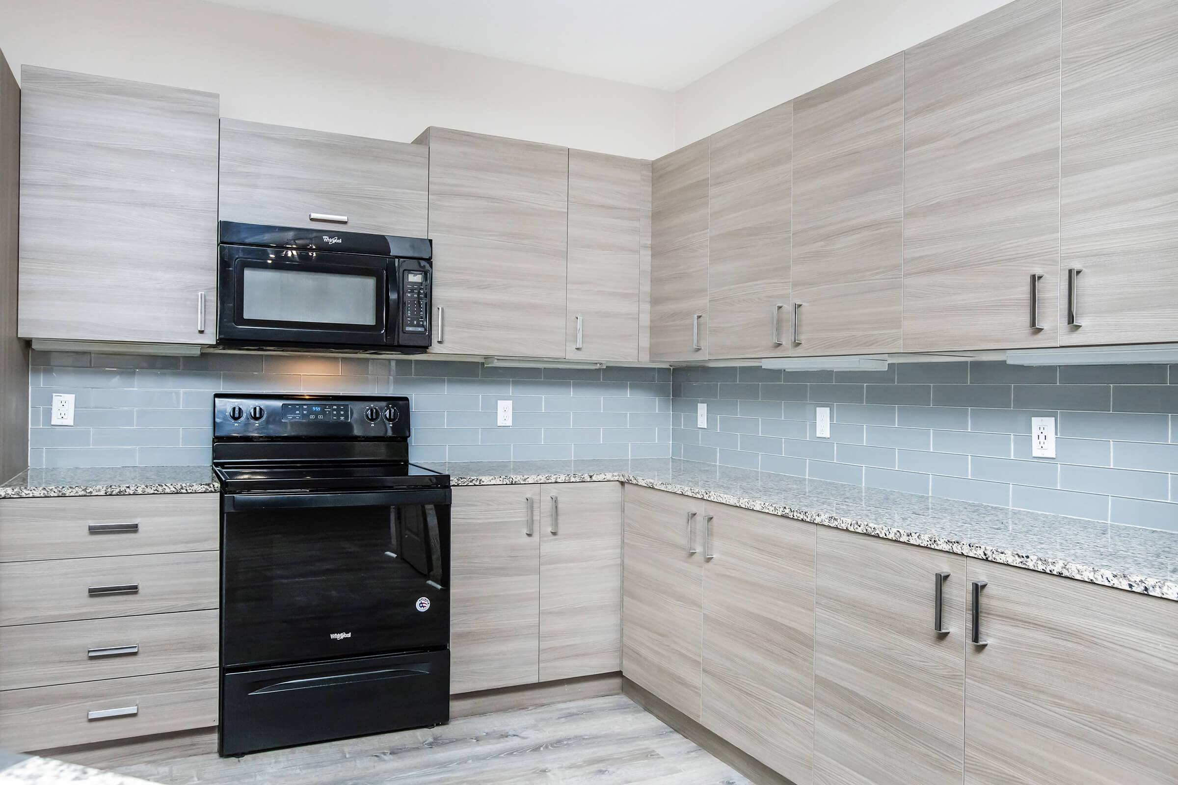 Modern kitchen featuring sleek, gray cabinetry and a granite countertop. The appliances include a black microwave and stove. The backsplash consists of light blue tiles, complementing the overall design. The space is bright and inviting, emphasizing a contemporary aesthetic.