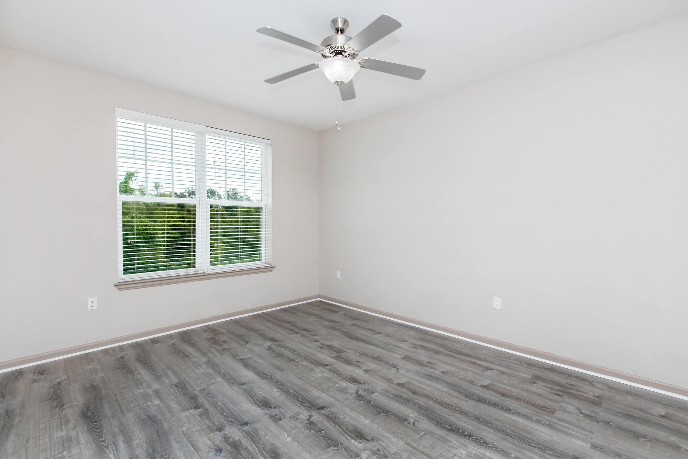 A bright, empty room with light gray walls and wooden flooring. There is a ceiling fan with five blades and a large window with white blinds, allowing natural light to illuminate the space. The view outside the window shows greenery, enhancing the room's airy feel.