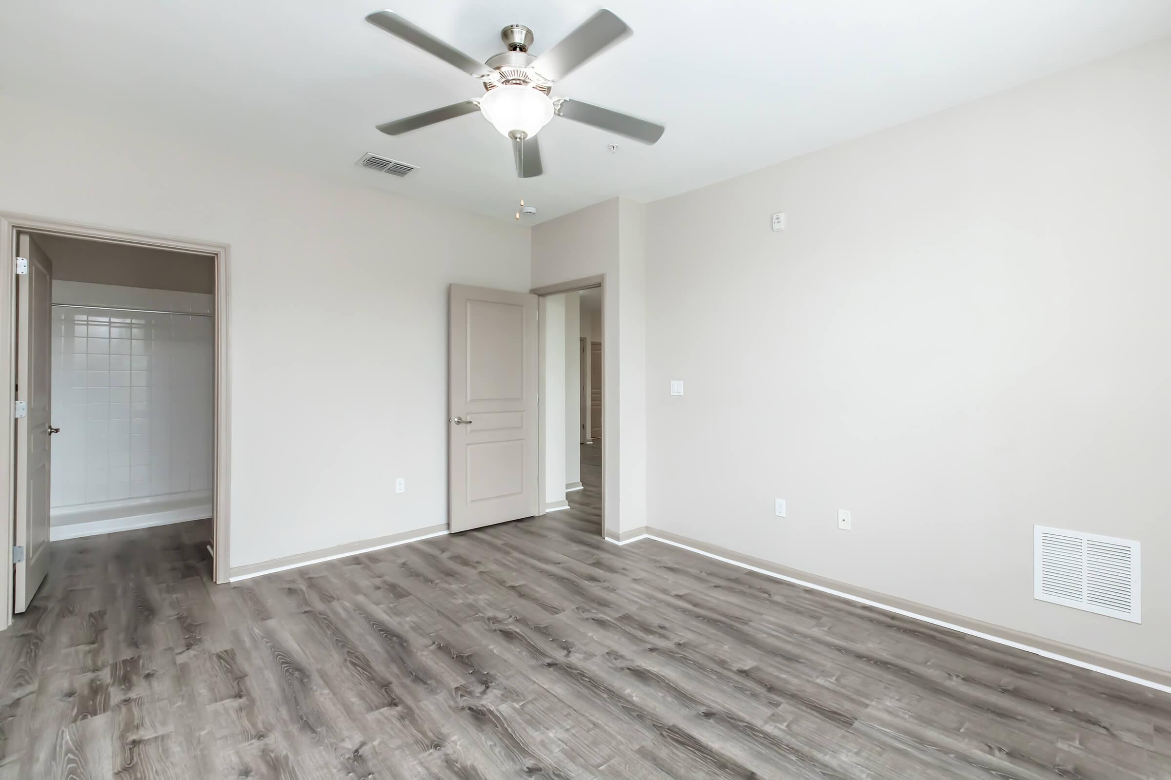 A spacious room with light gray walls and wooden flooring. It features a ceiling fan, a door leading to a bathroom area with a shower, and another door that appears to lead to another room. Natural light enhances the bright and open atmosphere of the space.