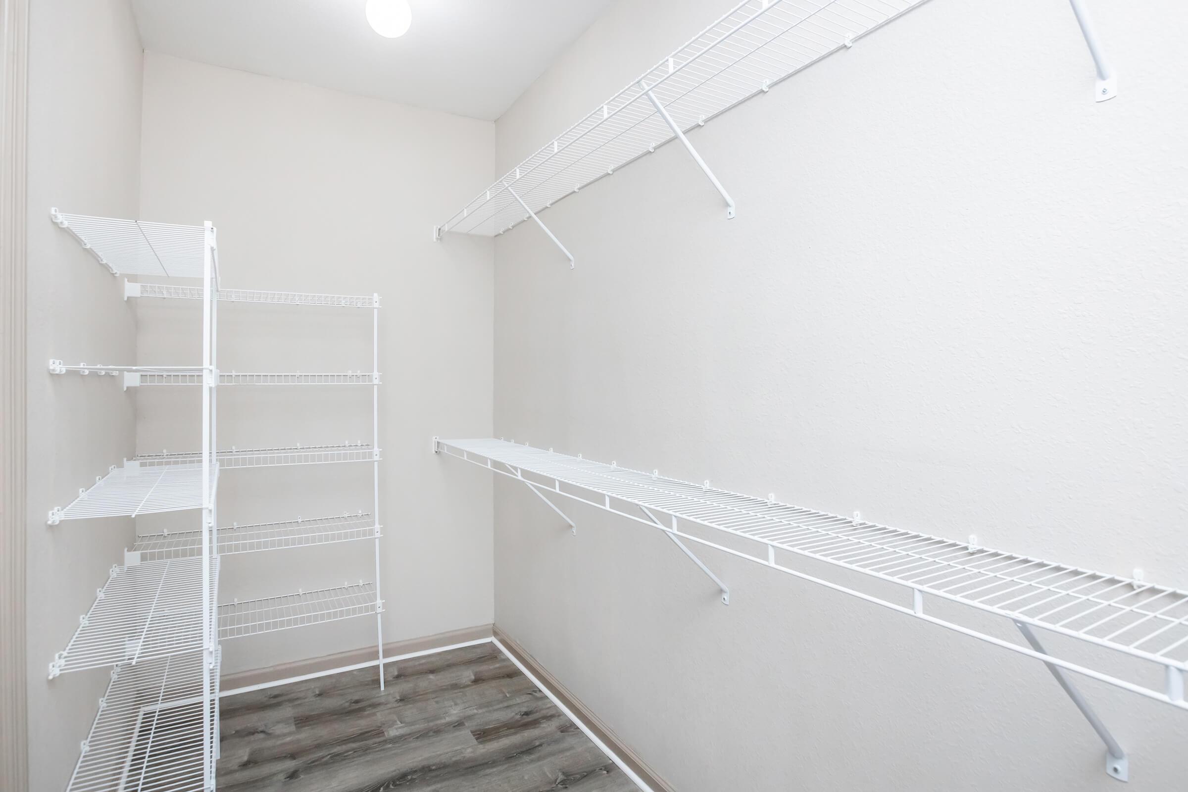 A clean and empty storage room featuring white wire shelves mounted on light beige walls. The floor has a wood-like appearance, and there is a ceiling light providing illumination. The shelves are arranged on opposite walls, creating an organized space for storage.