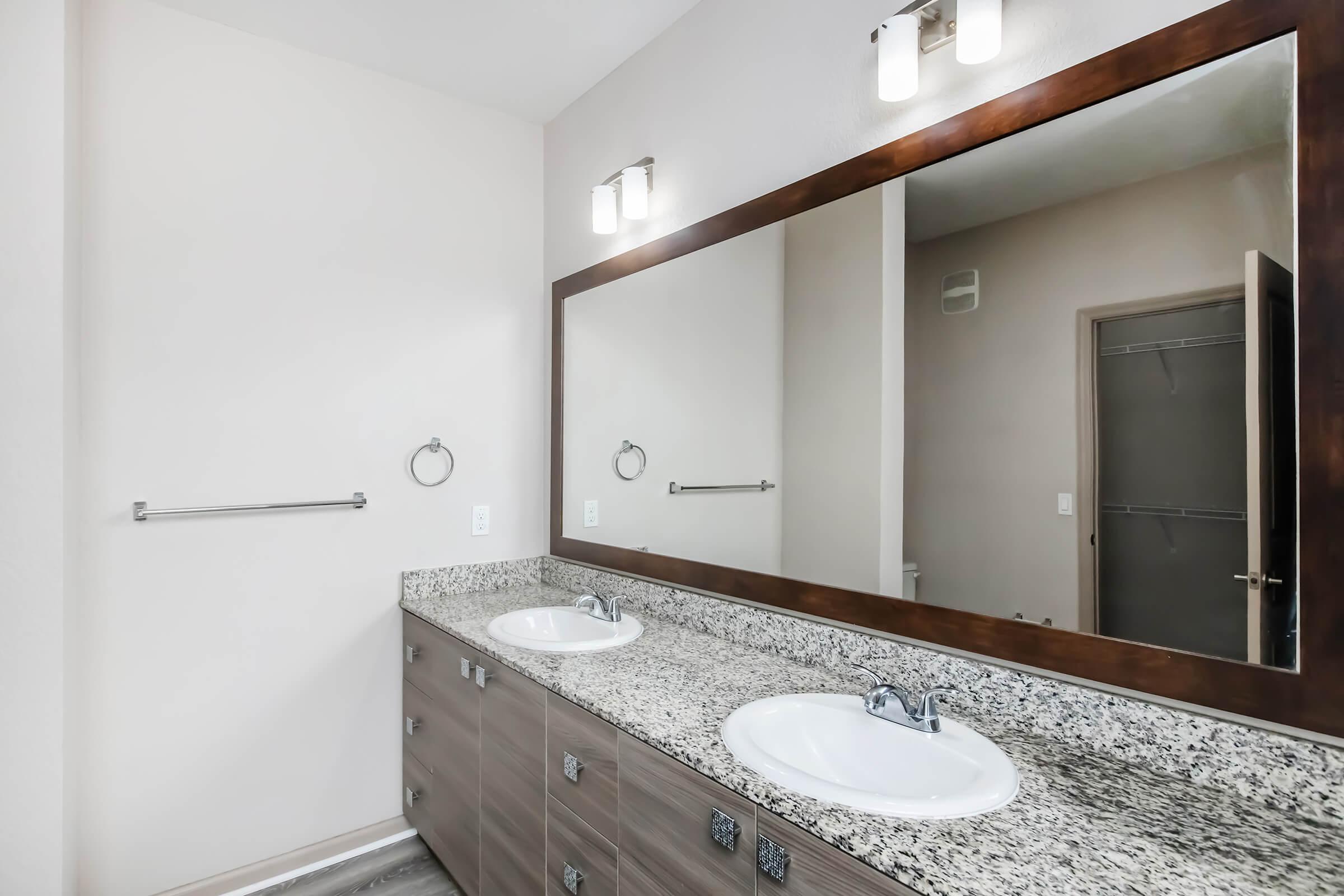 A modern bathroom with a large mirror above a double sink countertop made of speckled granite. The cabinetry below features sleek wooden finish, and there are two light fixtures above the mirror. A towel ring is mounted on the wall, and a closet door is visible in the background. The color scheme is neutral and bright.
