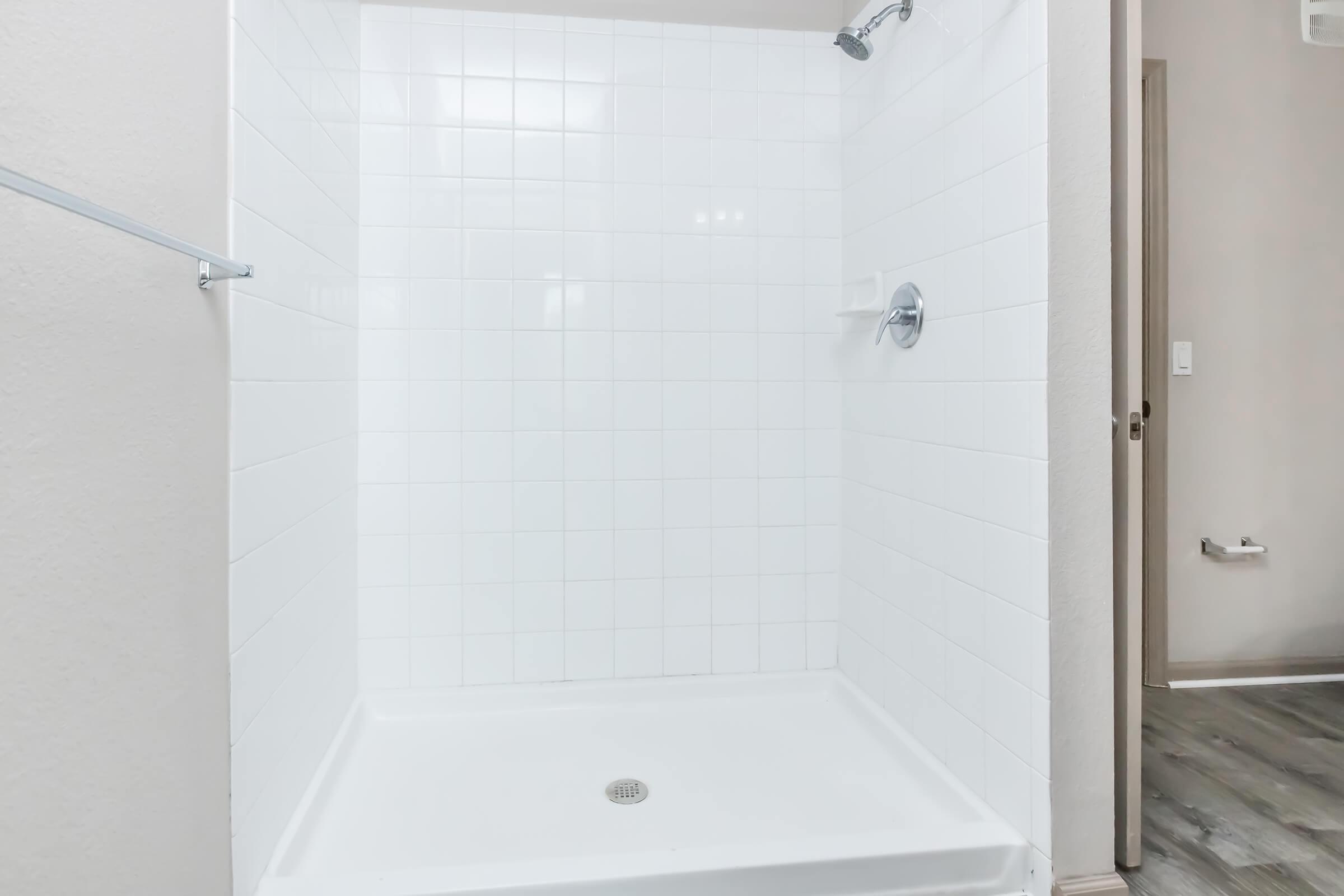 A clean, empty shower stall featuring white tiled walls and a modern showerhead. The floor is a white shower tray with a central drain, and there is a towel bar on one side. The lighting is bright, illuminating the minimalist design of the space.