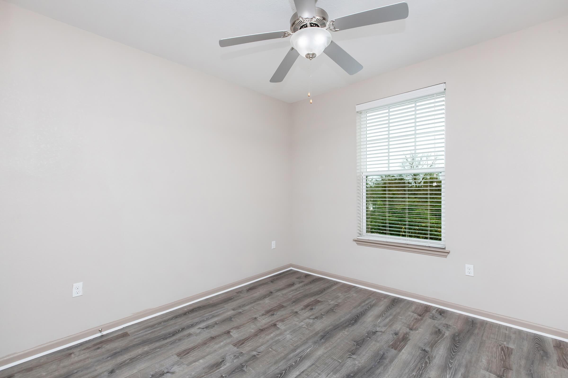 Empty room with light gray walls and a ceiling fan. A window with white blinds allows natural light to enter, revealing a view of greenery outside. The floor features wood-like laminate flooring. The room is unfurnished, creating a spacious and airy atmosphere.