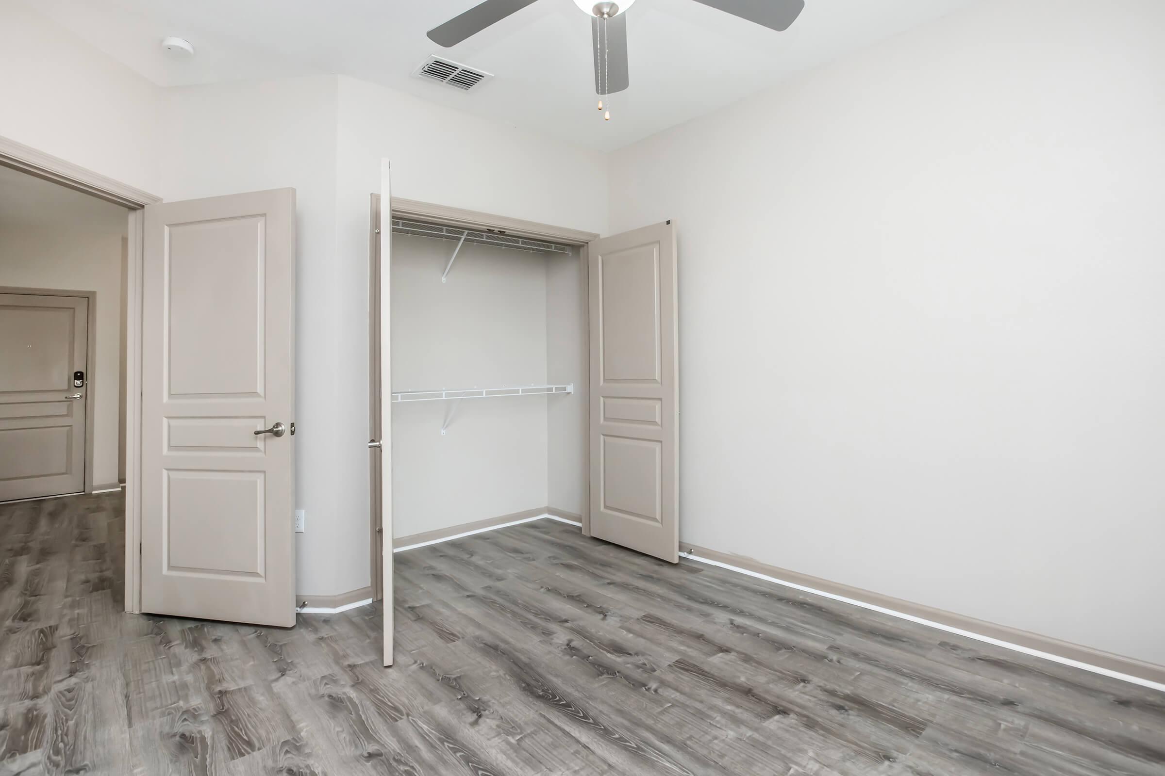 A spacious empty room with light-colored walls and a wooden floor. There is an open closet on the left with sliding doors, and a ceiling fan hangs from the ceiling. The room appears well-lit and ready for furniture.