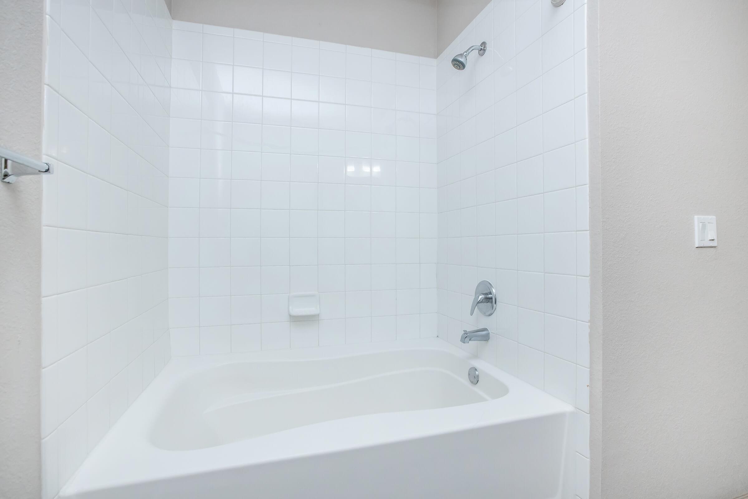 A clean and modern bathroom featuring a white tile shower area with a glass shower head, and a sleek, white bathtub. The walls are light-colored, enhancing the spacious feel. Natural light reflects off the tiles, creating a bright atmosphere.
