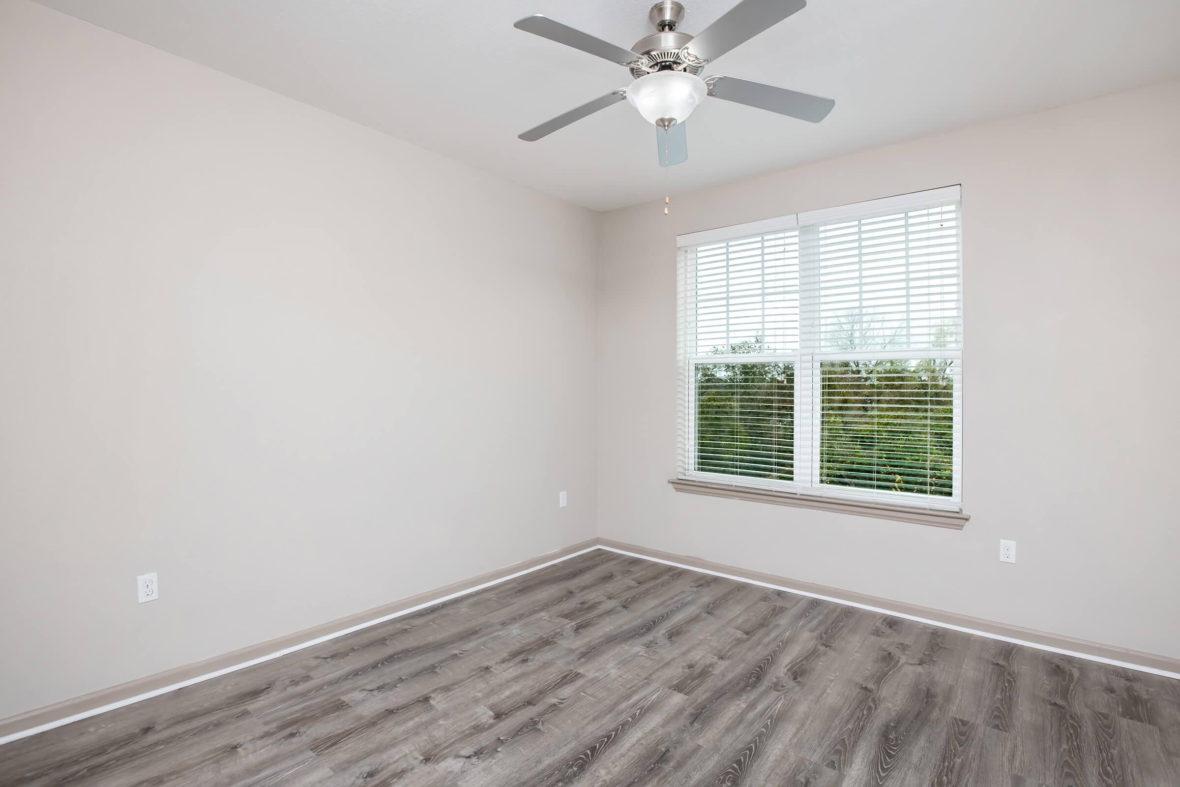 Empty room with light beige walls and a ceiling fan. A large window with white blinds lets in natural light, revealing a view of greenery outside. The flooring is laminate in a wood-like pattern, and the space is uncluttered, creating a bright and airy atmosphere.