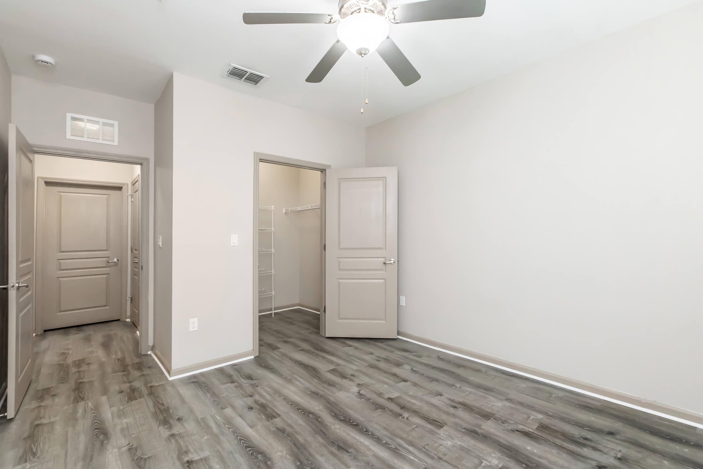 A well-lit, empty room with light gray walls and laminate wood flooring. A ceiling fan hangs in the center, and there are two doorways: one leading to a closet and another to an adjoining space. The overall ambiance is modern and minimalistic.