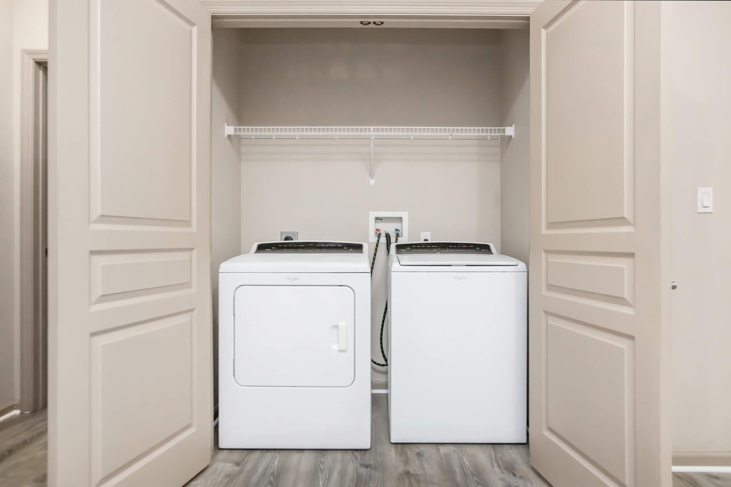 A laundry room featuring a side-by-side washer and dryer set in a light-colored space with beige walls. Above the appliances, there is a shelf for storage. The room has double doors that open into the laundry area, with a neutral flooring design.