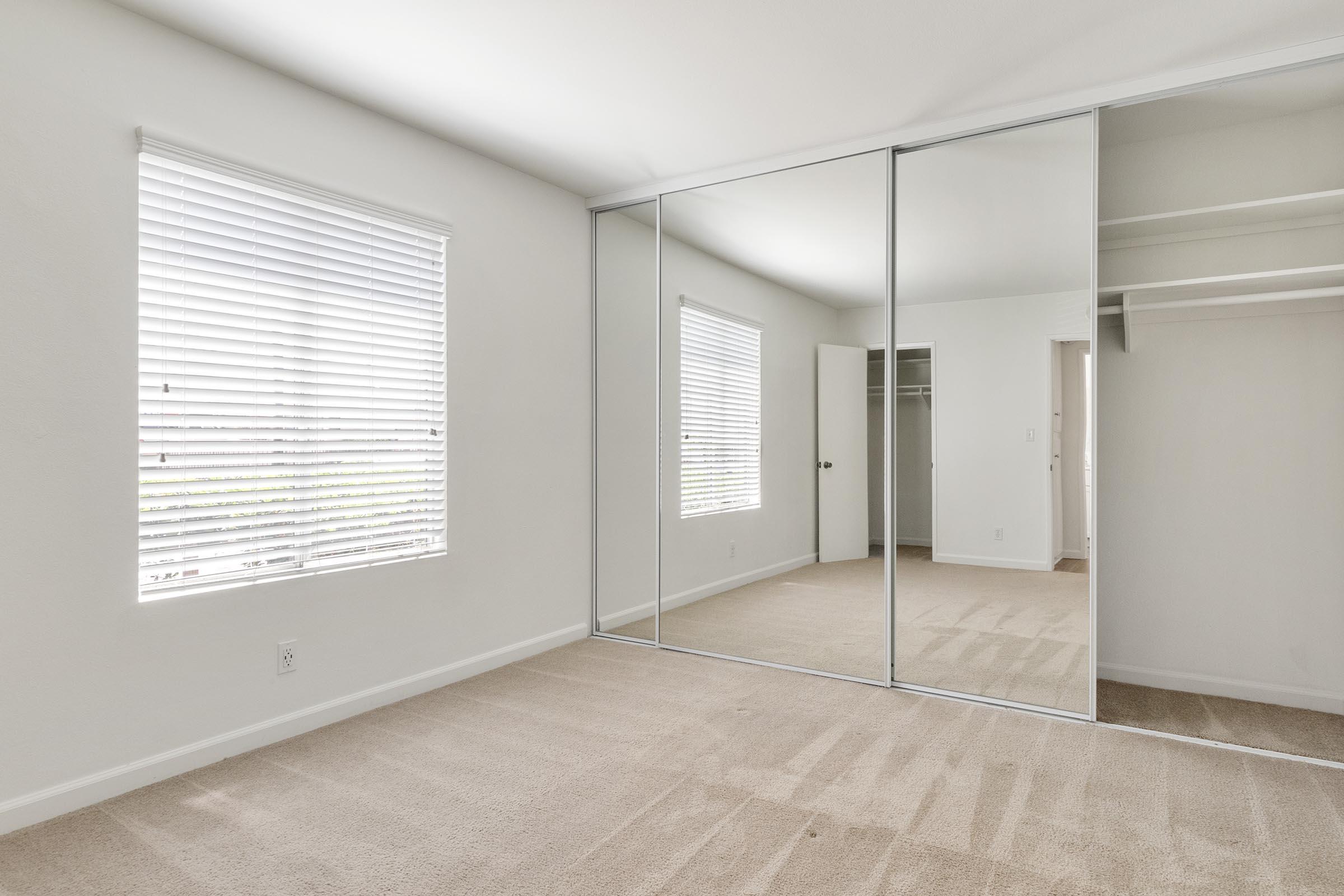 Empty room with light-colored walls and beige carpet. A large mirrored closet spans one wall, reflecting natural light from a nearby window with white blinds. The room is spacious and simple, featuring a door leading to another area and no furniture present.