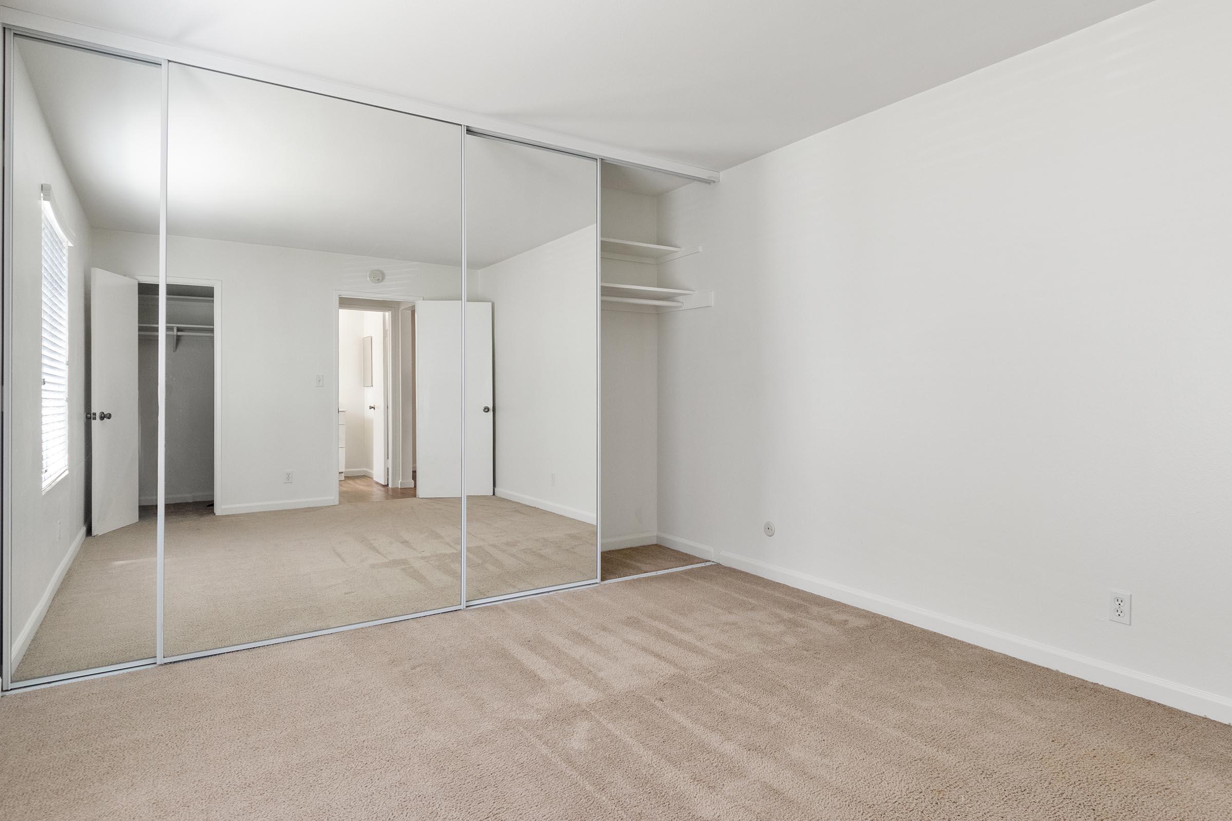 A spacious, empty room featuring a large mirror closet and soft beige carpet. There is natural light coming through a window, highlighting the clean, minimalist design. The room has white walls and a door leading to another space, suggesting a well-maintained living area.