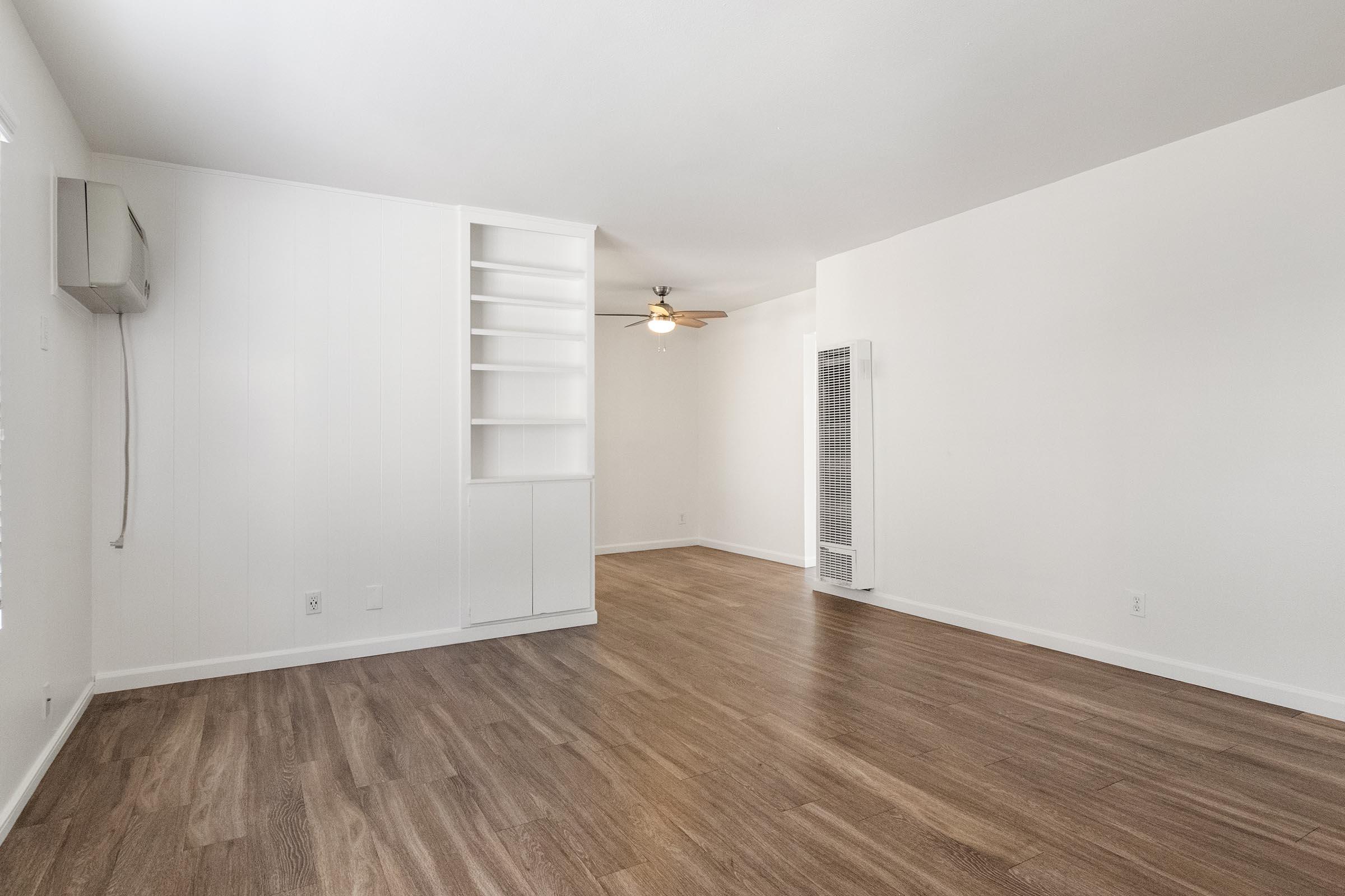 A spacious, empty room featuring light-colored walls and wooden flooring. It includes a ceiling fan, a built-in shelving unit on one wall, and an air conditioning unit. Natural light enters through a window, giving the room a bright appearance. The layout is open, creating a sense of airy simplicity.