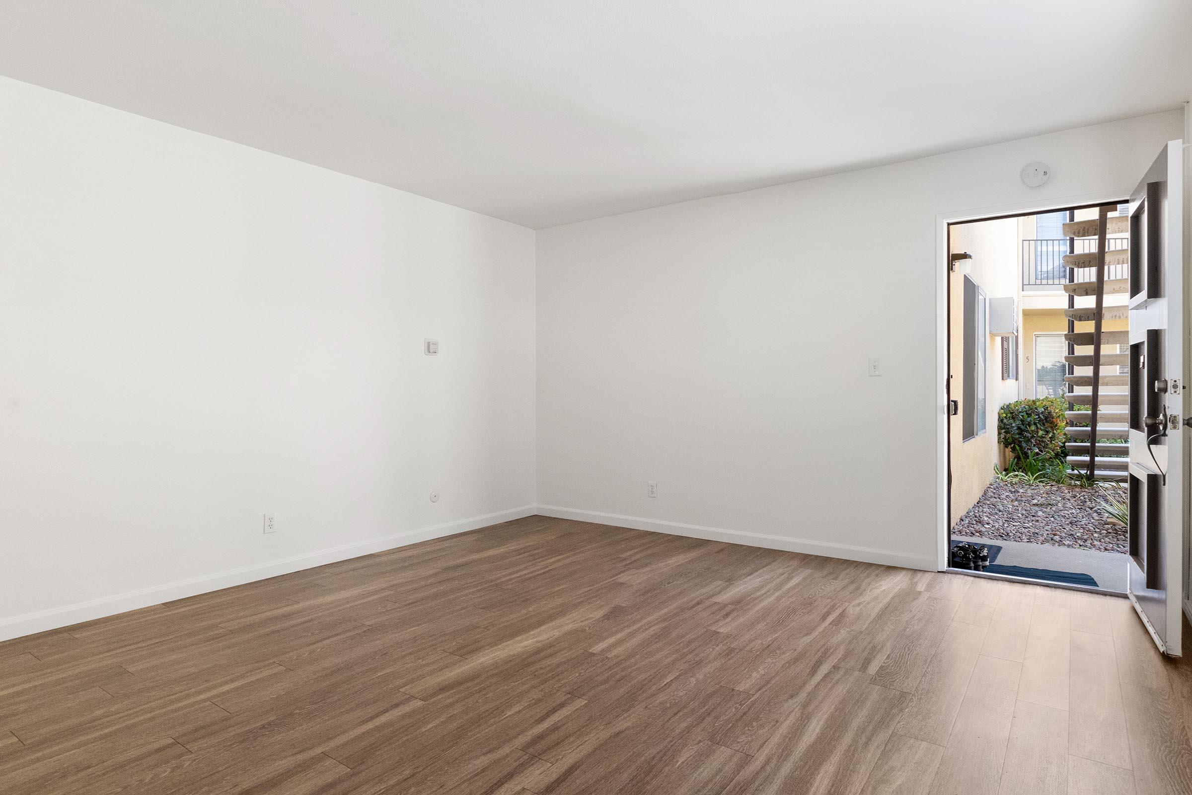 Empty interior of a room with light-colored walls and wood flooring. An open door leads to an outdoor area with a staircase and greenery visible outside. Natural light brightens the space, emphasizing its simplicity and spaciousness. No furniture is present.