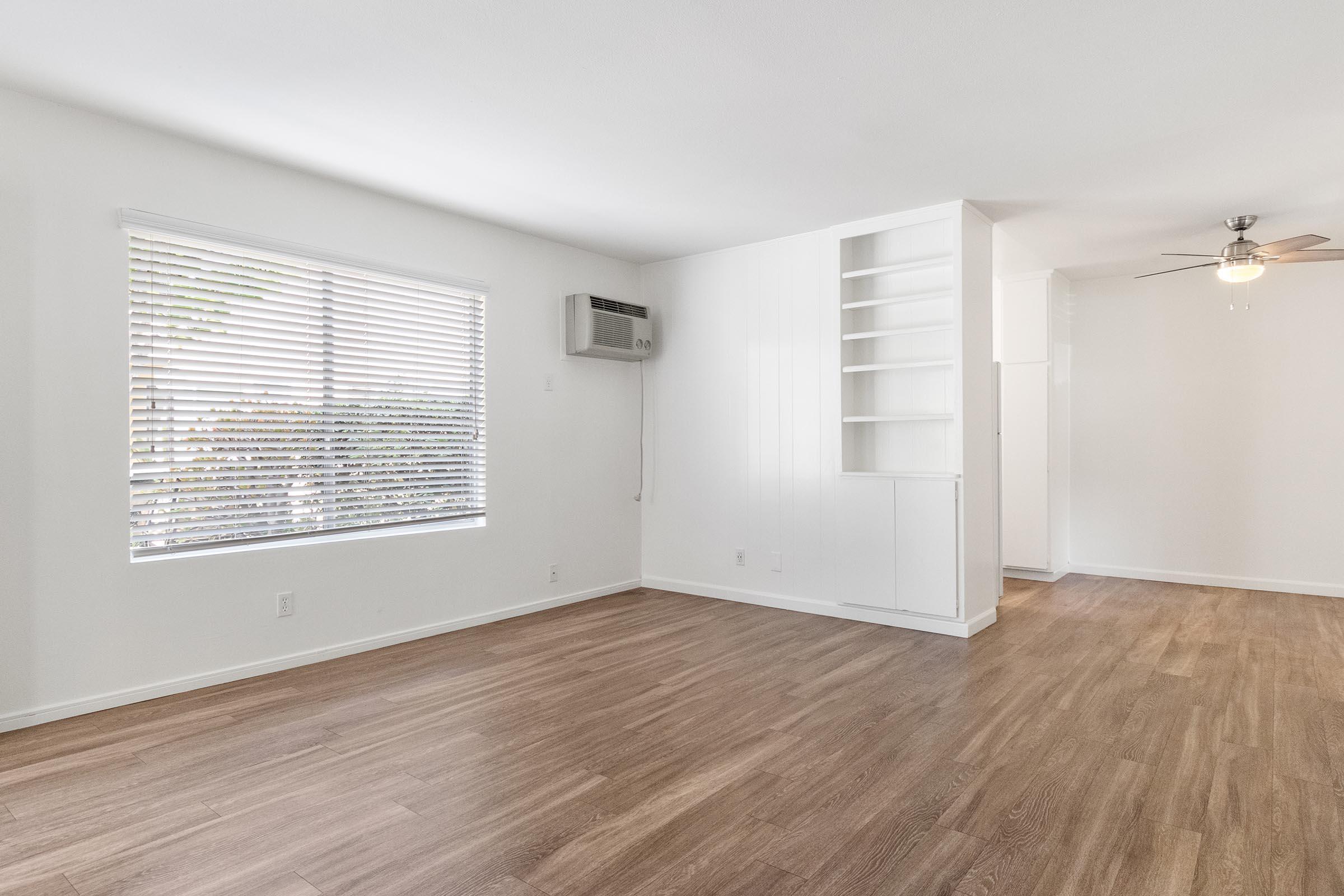A bright, spacious room with large windows showcasing blinds, an air conditioning unit on the wall, and wood-look flooring. The room features a light color scheme and a ceiling fan. There is a nearby nook with built-in shelves, creating an open and inviting living space.