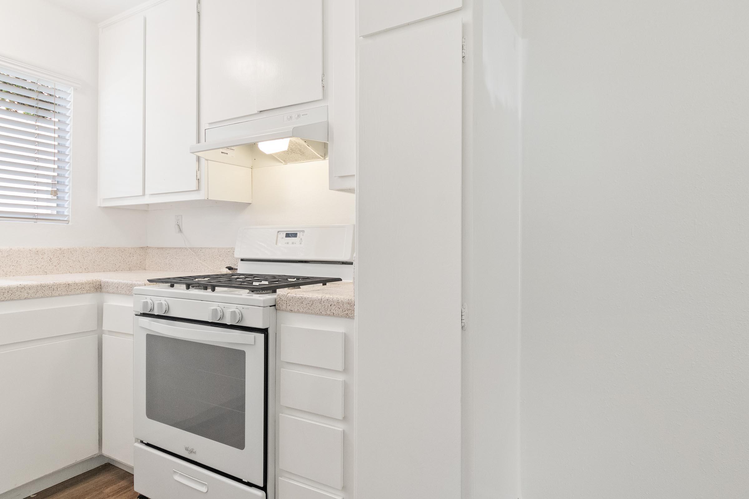 A bright kitchen featuring white cabinets and a light-colored countertop. A modern gas stove is visible with a range hood above it, and there are several drawers and a window with blinds, allowing natural light to illuminate the space. The flooring is a warm wood tone, adding a cozy feel.