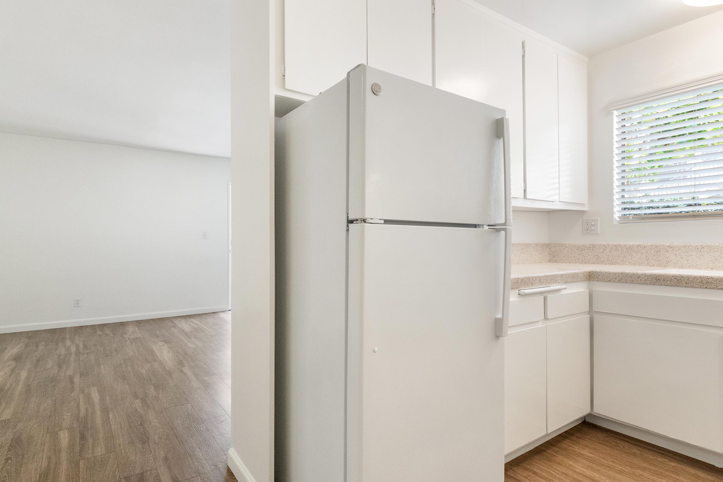 A modern kitchen featuring a white refrigerator, light-colored cabinets, and a countertop. There is a window with blinds allowing natural light, and wood-look flooring is visible. An adjacent living space can be seen in the background.