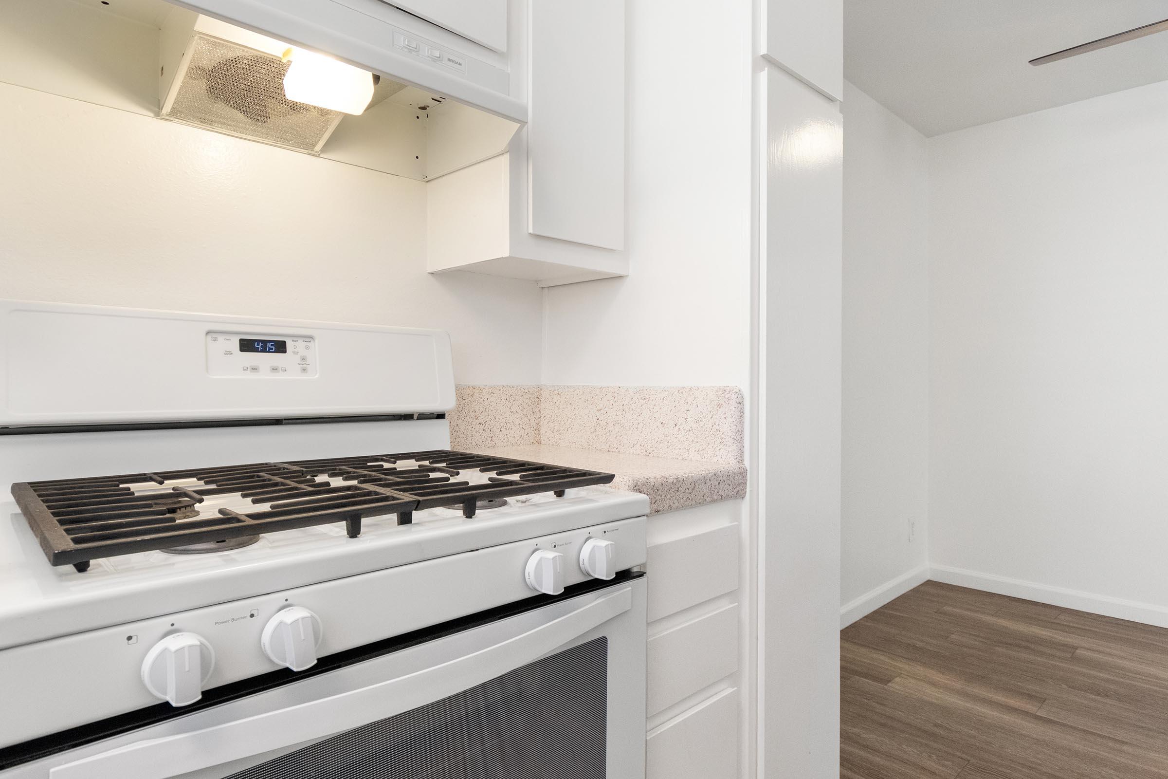 A modern kitchen featuring a white gas stove with a five-burner cooktop, a standard oven below, and beige countertops. The background shows a bright, minimalistic space with light-colored walls and wooden flooring, suggesting a clean and inviting dining area.