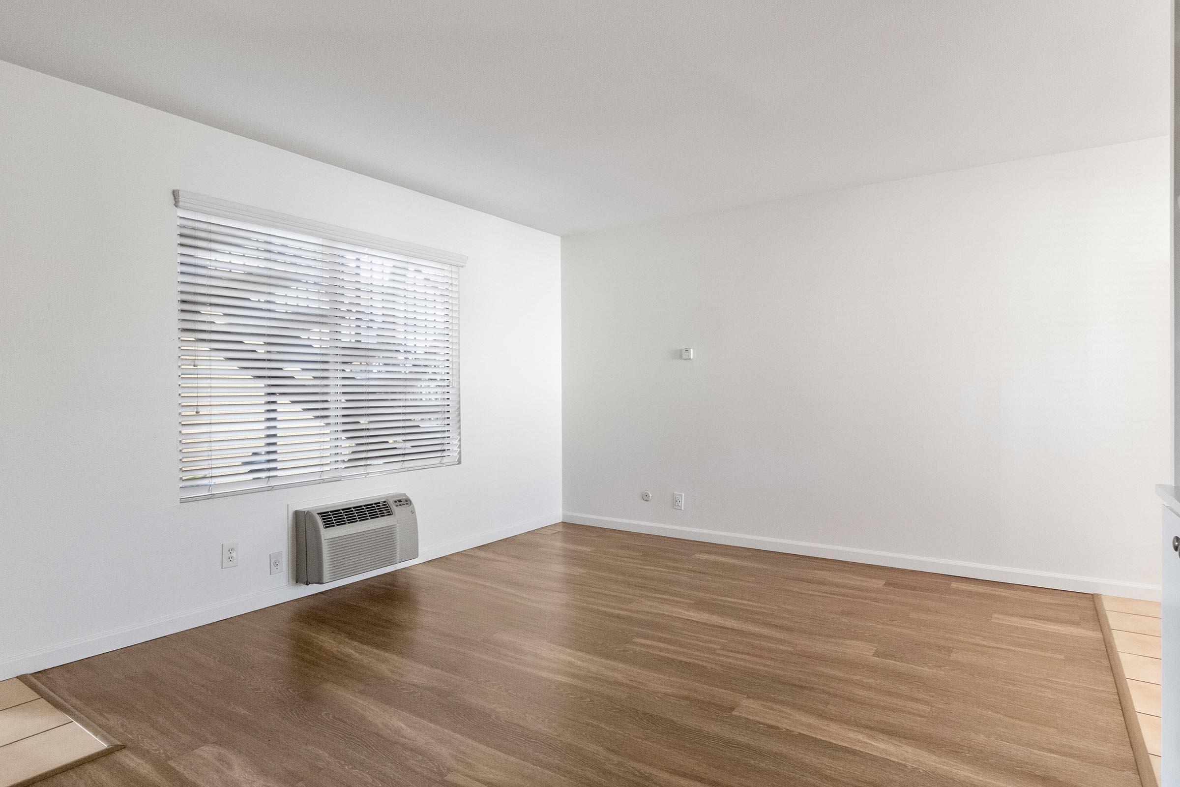 A bright, empty room with light-colored walls and a wooden floor. A window with white blinds lets in natural light, and there is an air conditioning unit on the floor near the wall. The space is minimalist and clean, with no furniture or decorations.